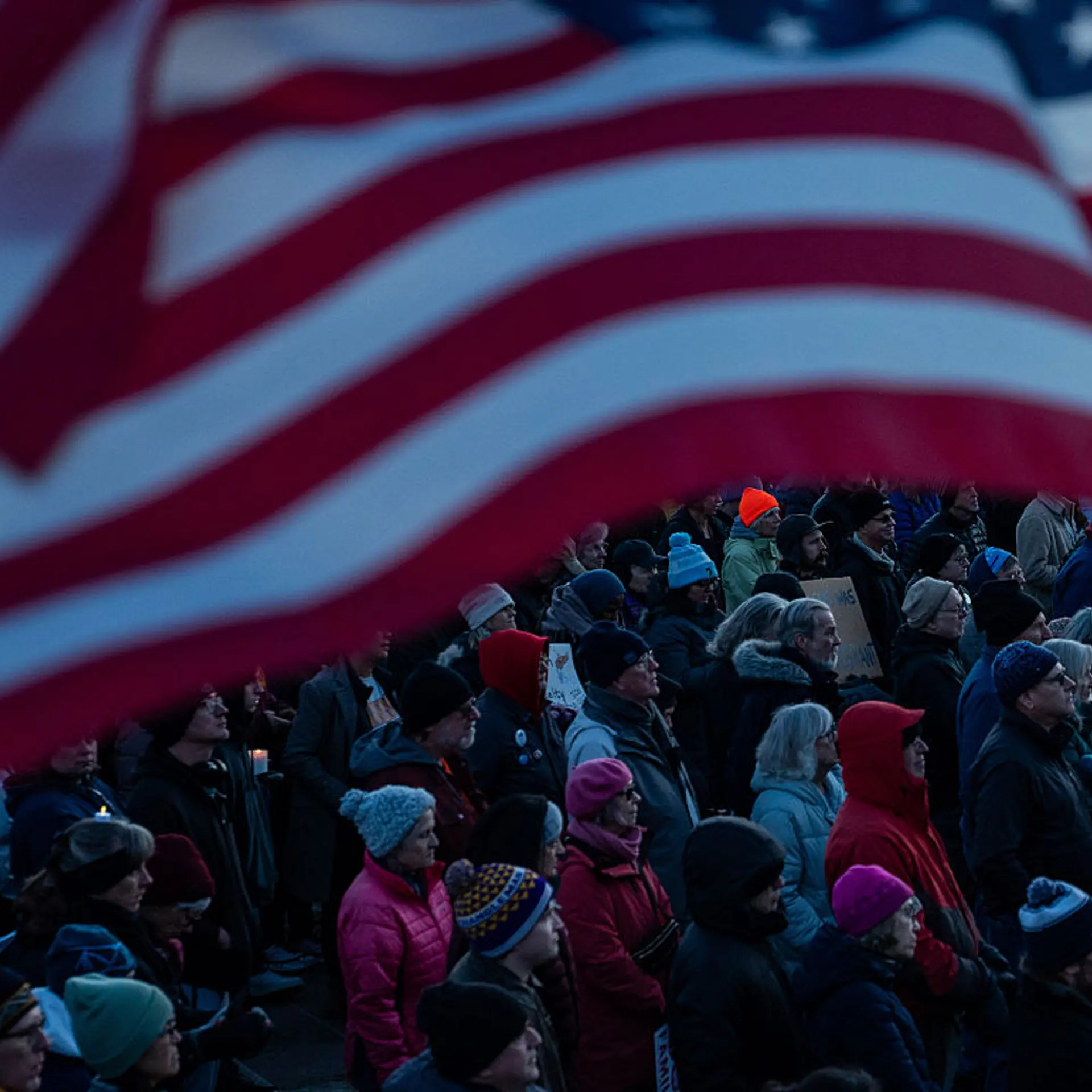 An American flag waves in front of a crowd of people gathered outside in silent vigil.