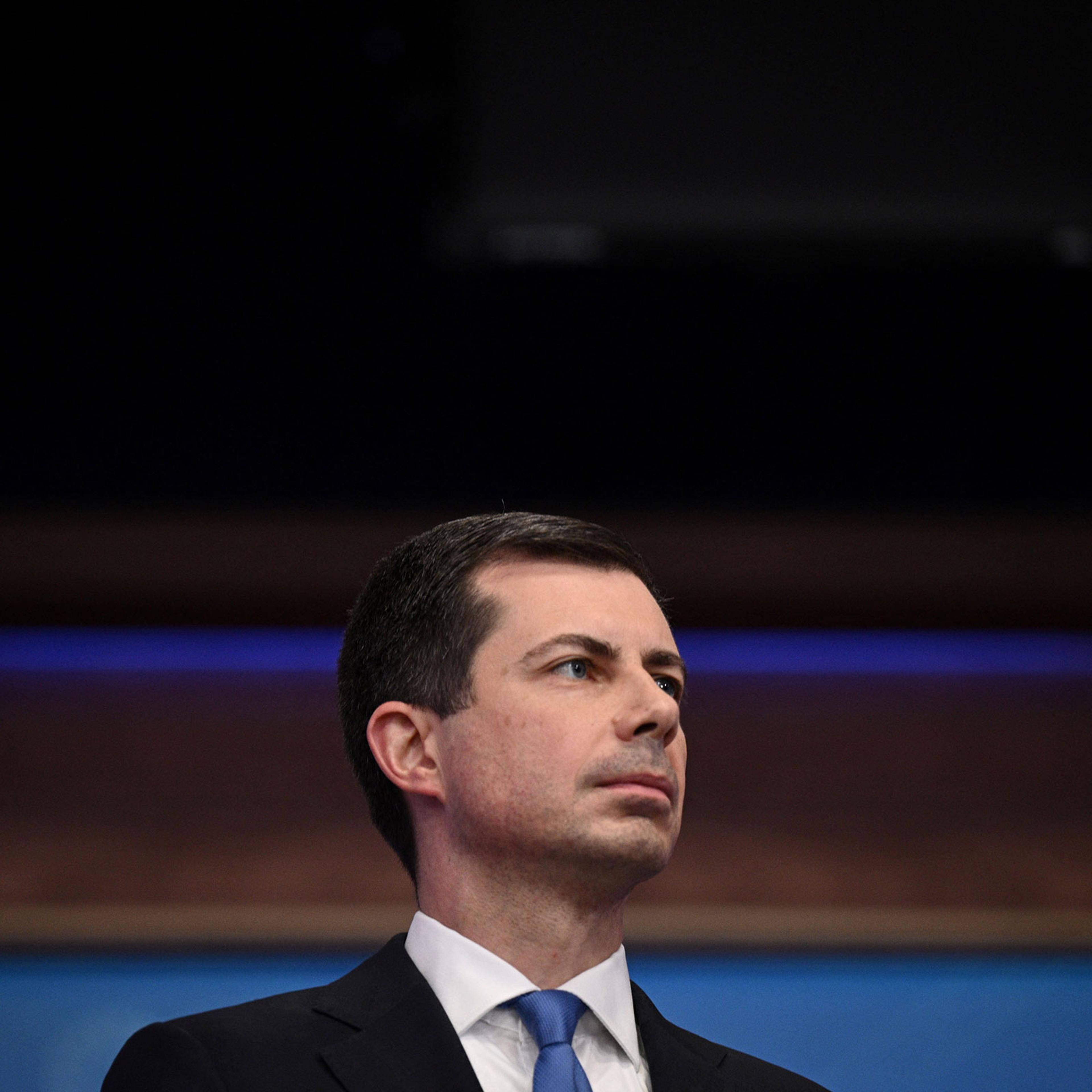 Transportation Secretary Pete Buttigieg in a suit with a white shirt and blue tie looks off to the side while delivering remarks during a press conference