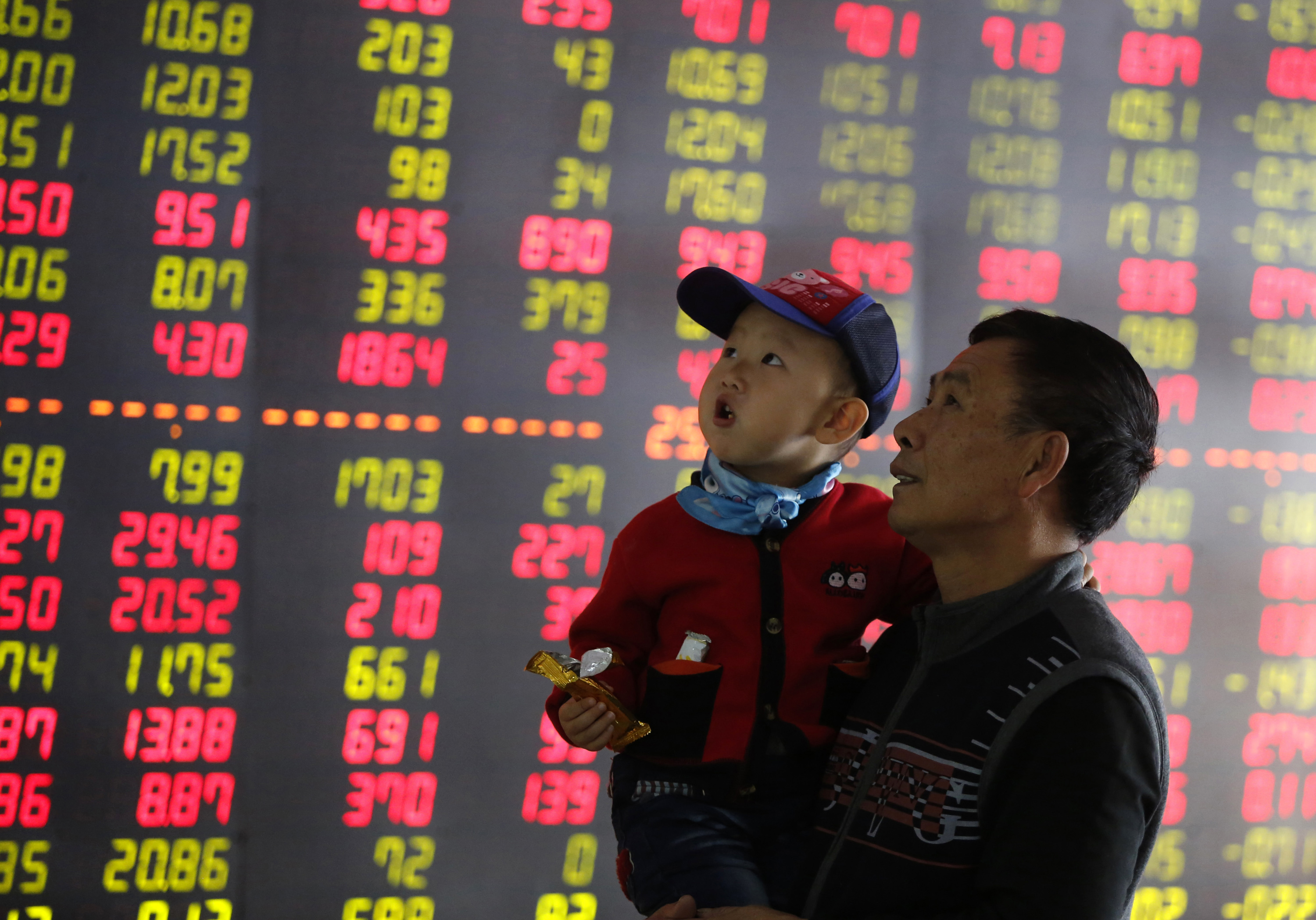 An investor holds a child in front of an electronic screen showing stock information at a brokerage house in Shenyang