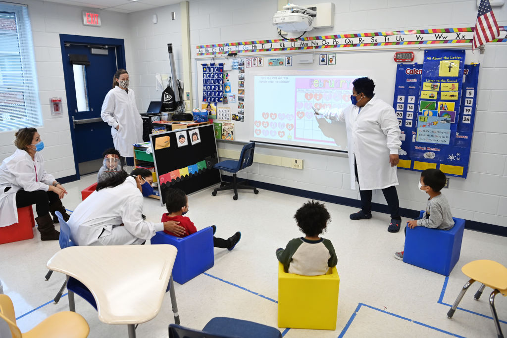 A teacher dressed up in a white lab coat and a mask teaches her preschool autism class with four students sitting in a semicircle around awhiteboard. There are three other teachers also dressed in white lab coats and masks—two are listening to the class 