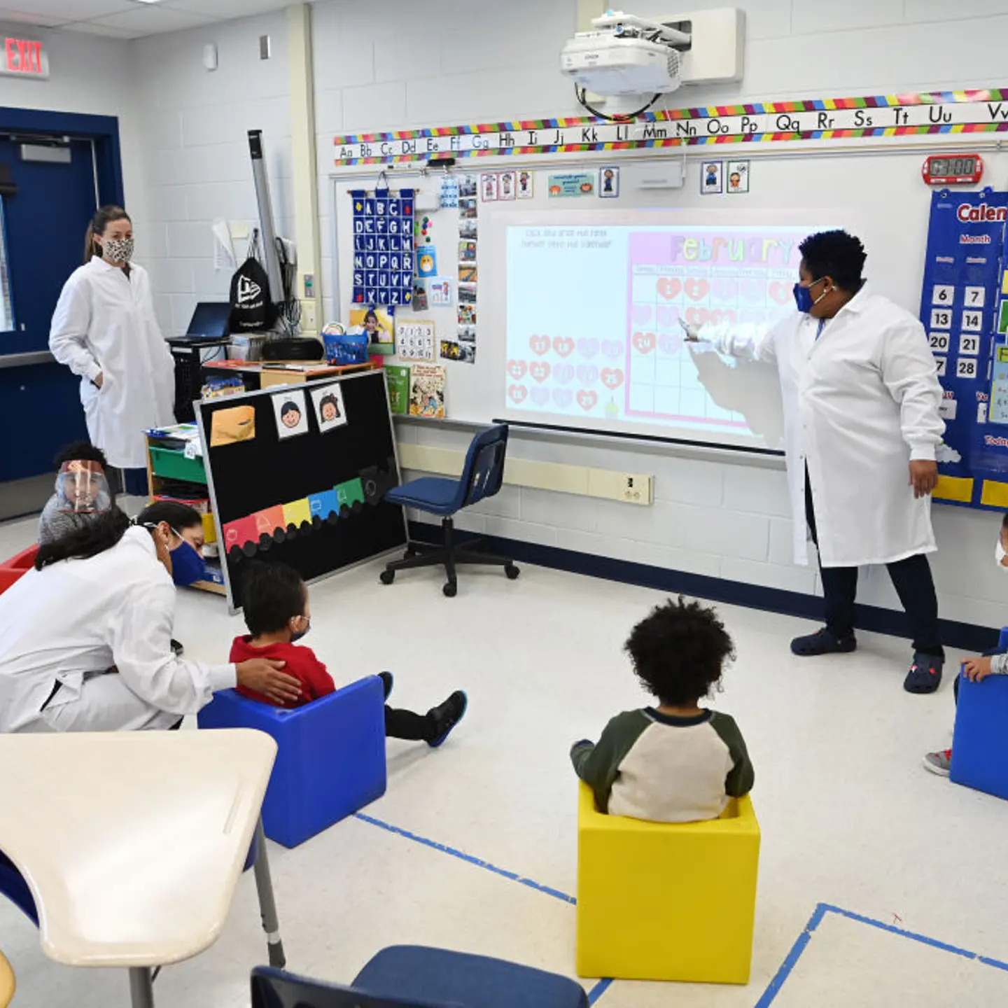 A teacher dressed up in a white lab coat and a mask teaches her preschool autism class with four students sitting in a semicircle around awhiteboard. There are three other teachers also dressed in white lab coats and masks—two are listening to the class
