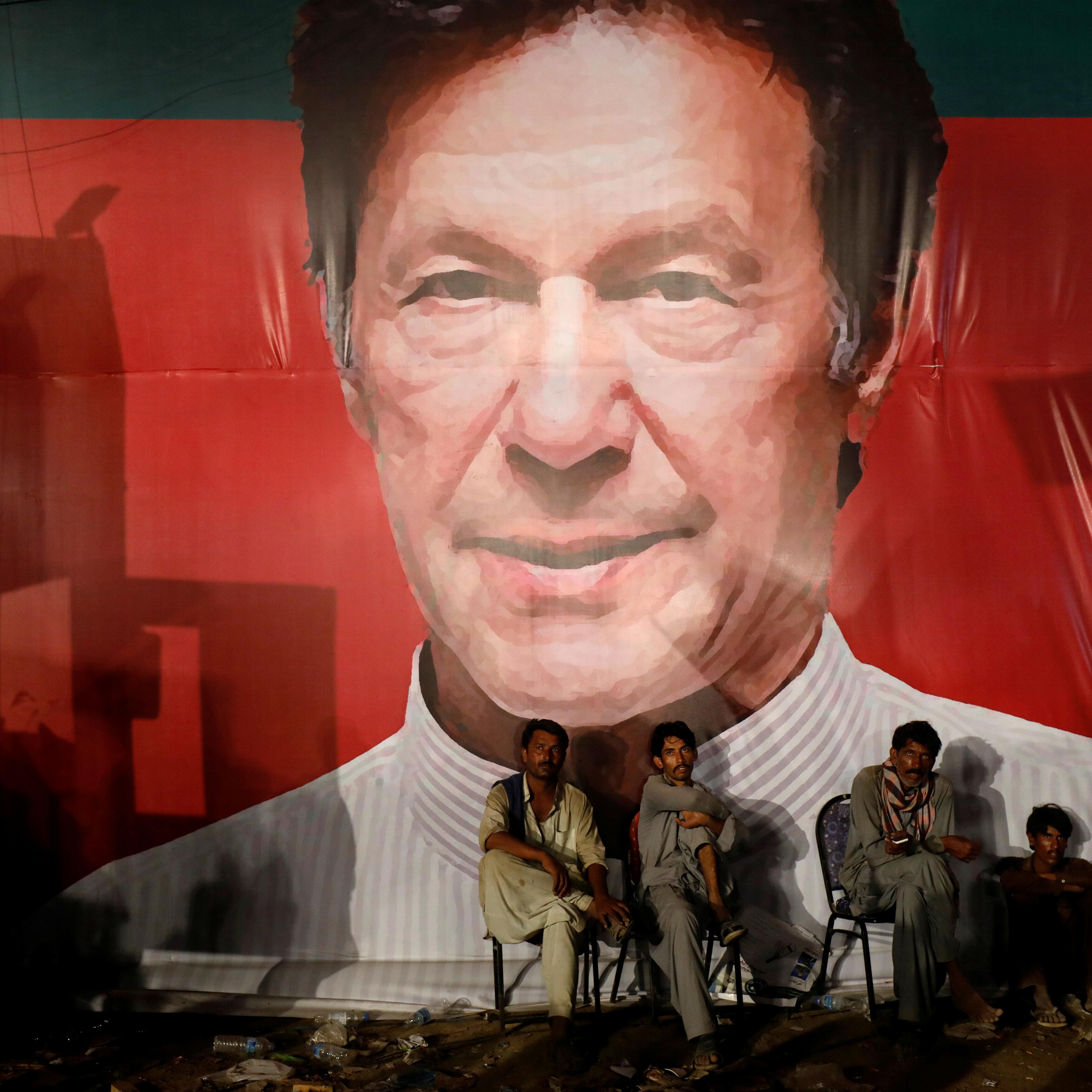 Labourers, who set up the venue, sit under a wall with a billboard displaying photo of Imran Khan, chairman of the Pakistan Tehreek-e-Insaf (PTI), political party, as they listen to him during a campaign rally ahead of general elections in Karachi