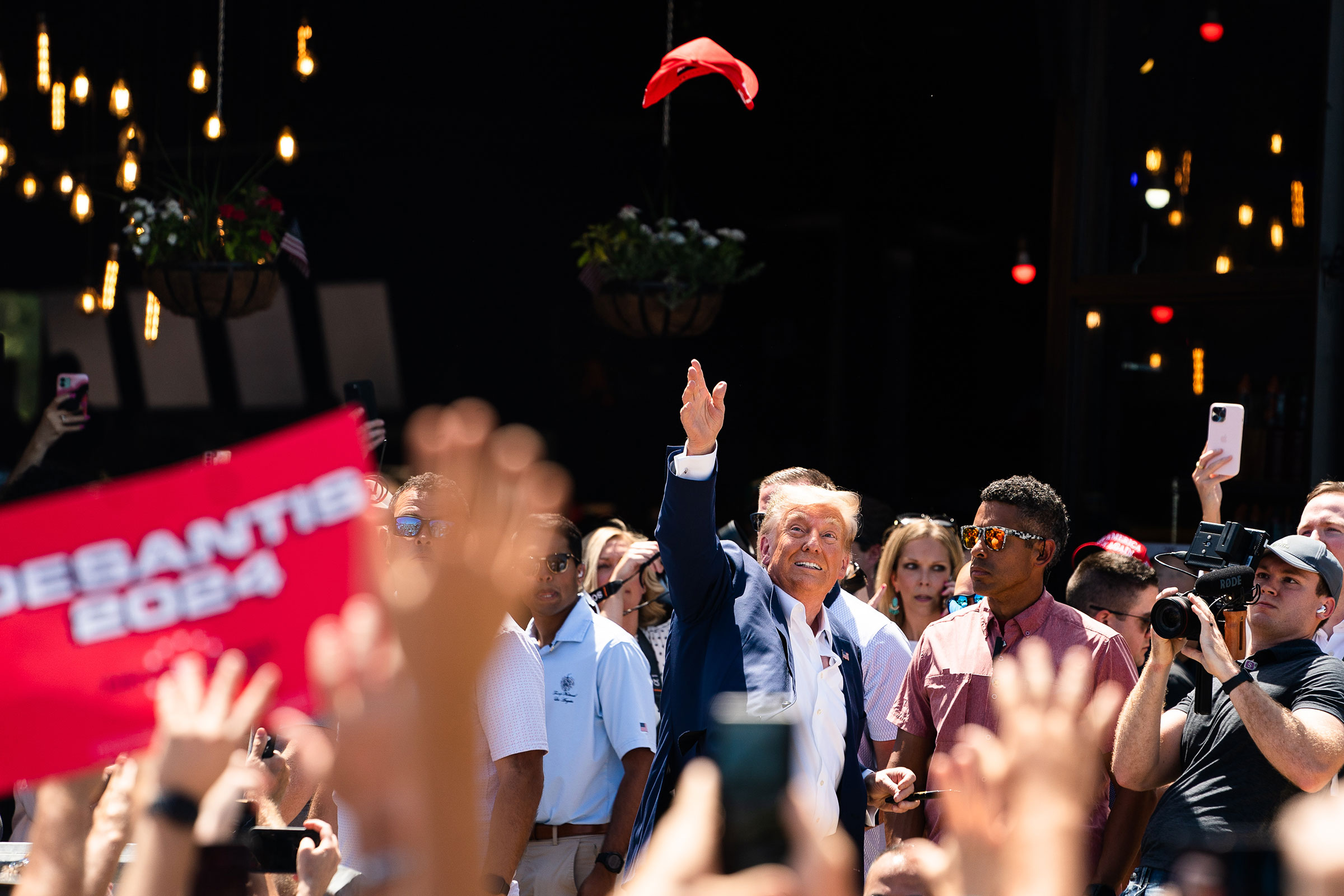 Donald Trump throws a red hat into the air as he is surrounded by reporters, staff, and supporters