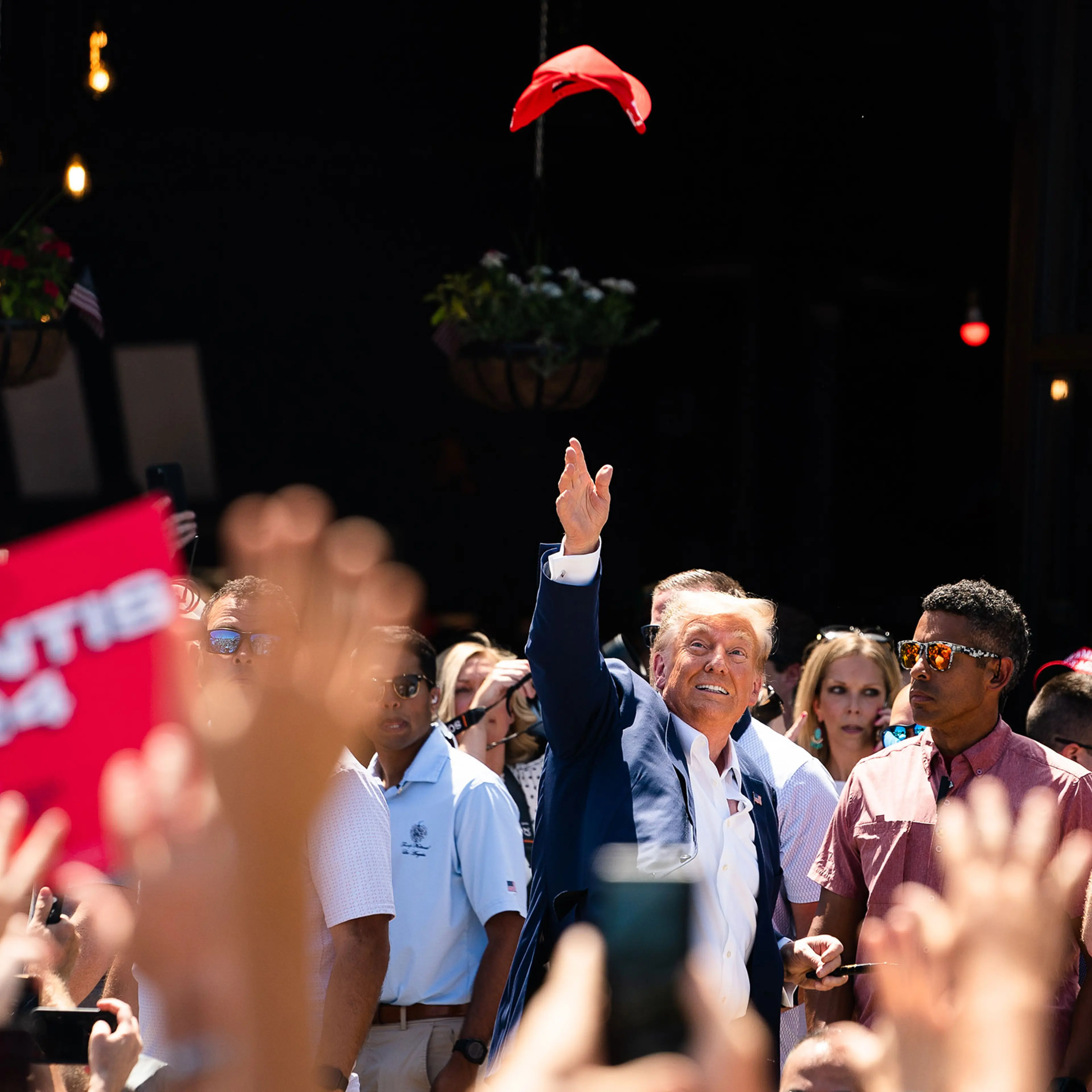 Donald Trump throws a red hat into the air as he is surrounded by reporters, staff, and supporters
