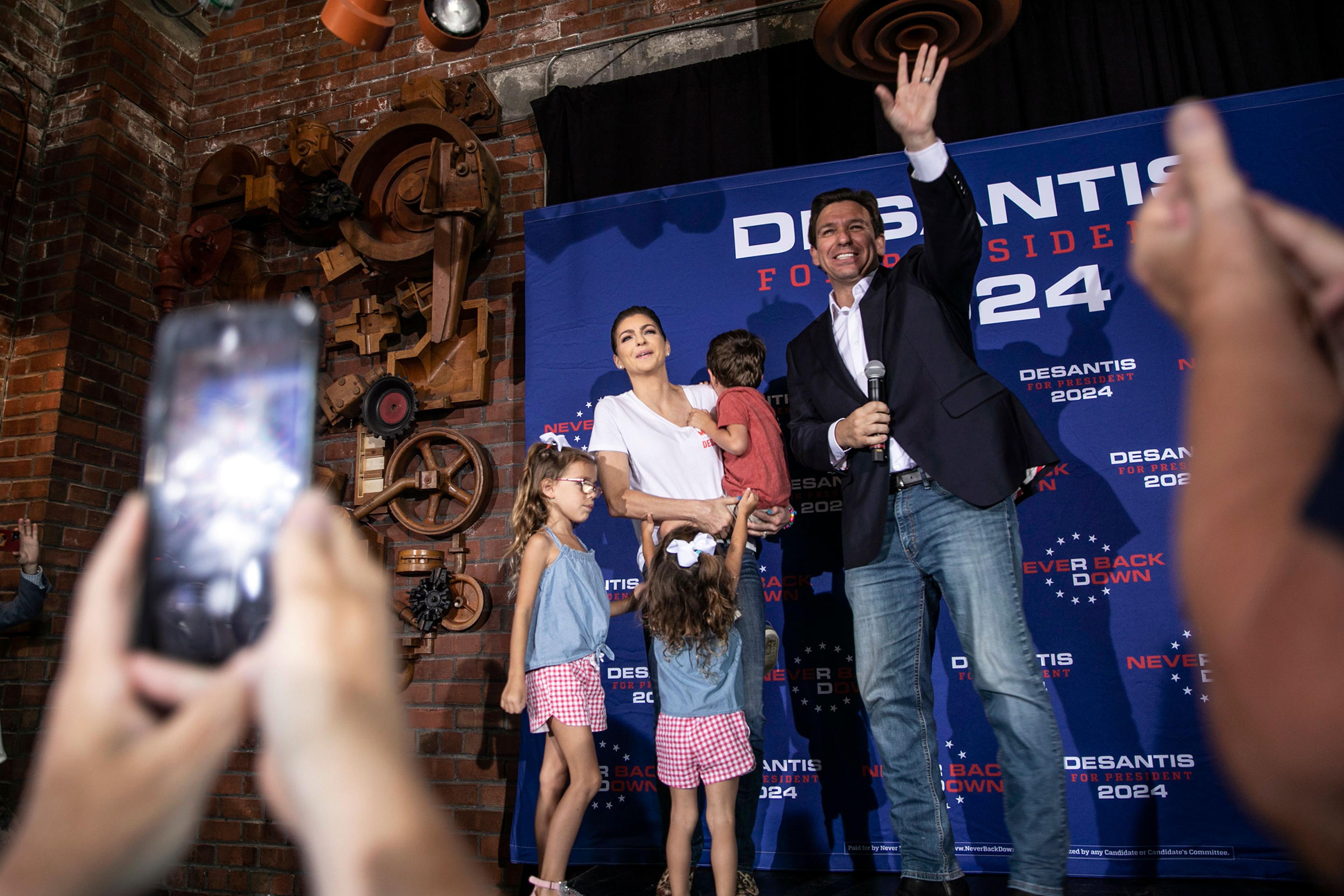 Ron DeSantis stands on a small stage and waves at supporters while his wife stands next to him with their three children