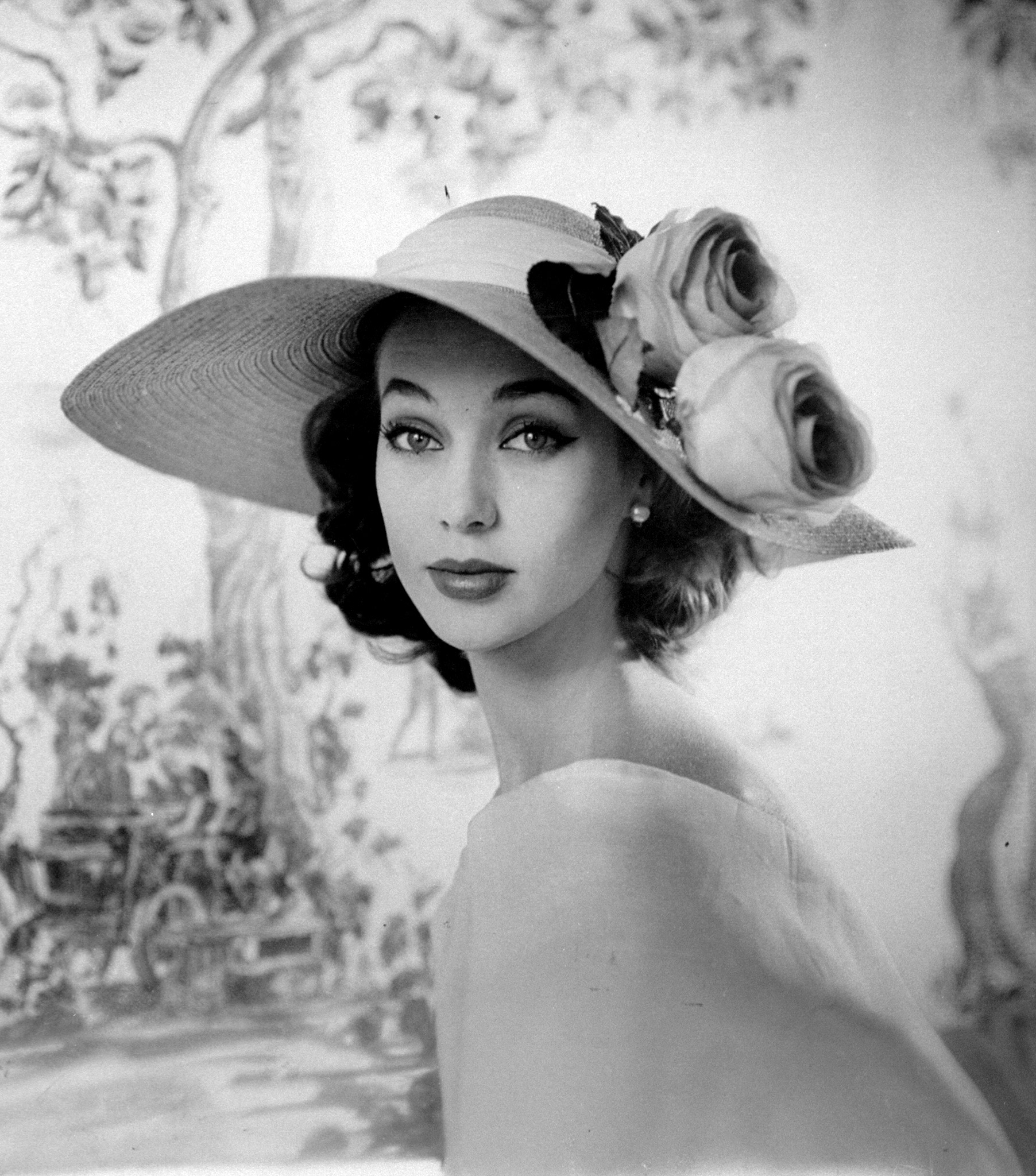 A woman wearing a party hat with two flowers decorated in the traditional garden style as part of the Gainsboro fashion collection. 1956.