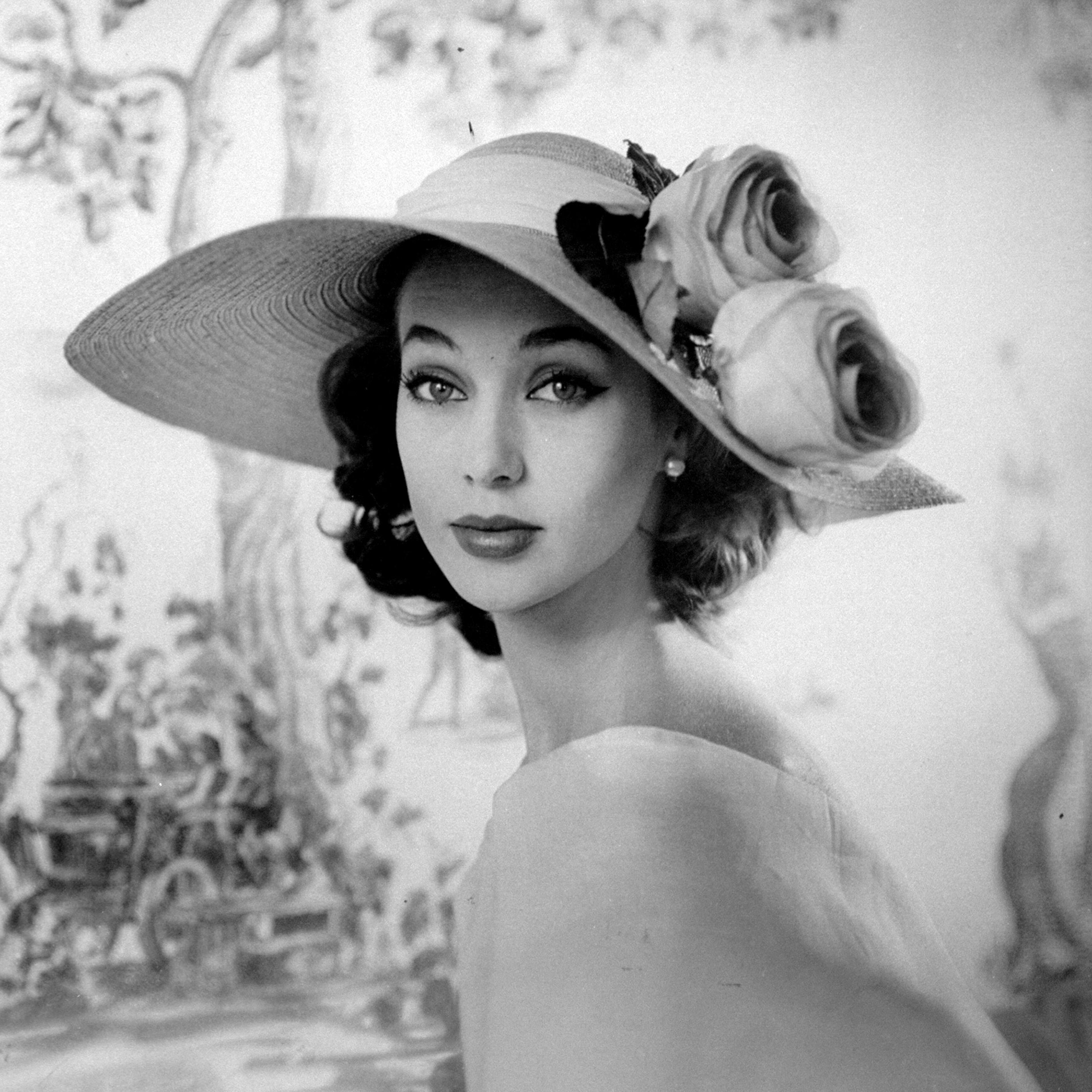 A woman wearing a party hat with two flowers decorated in the traditional garden style as part of the Gainsboro fashion collection. 1956.