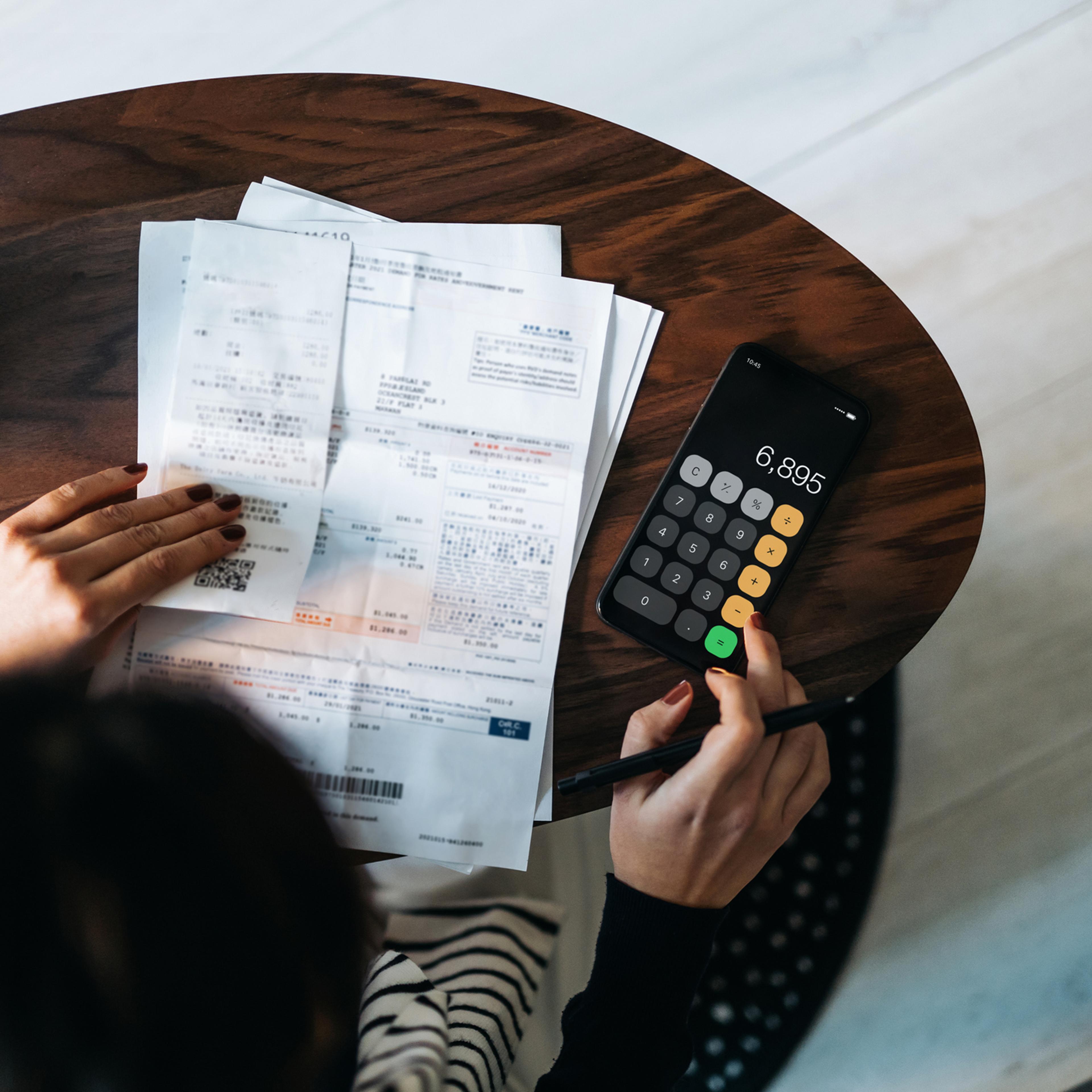 Overhead view of young Asian woman managing personal banking and finance at home. Planning budget and calculating expenses while checking her bills with calculator. Managing taxes and financial bills. Home budgeting. Concept of finance and economy
