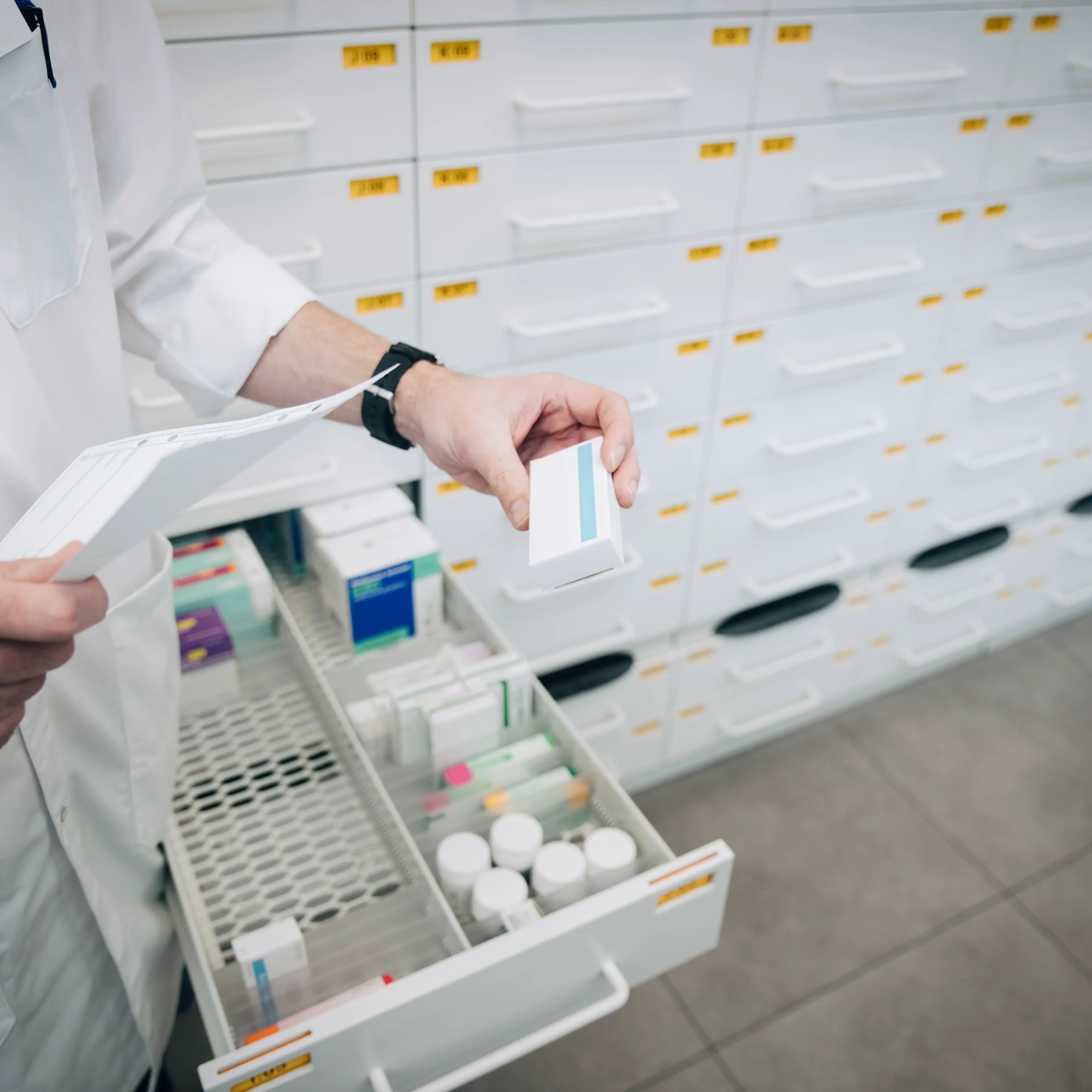 Midsection of mature male pharmacist holding prescription paper and medicine standing by drawer at store