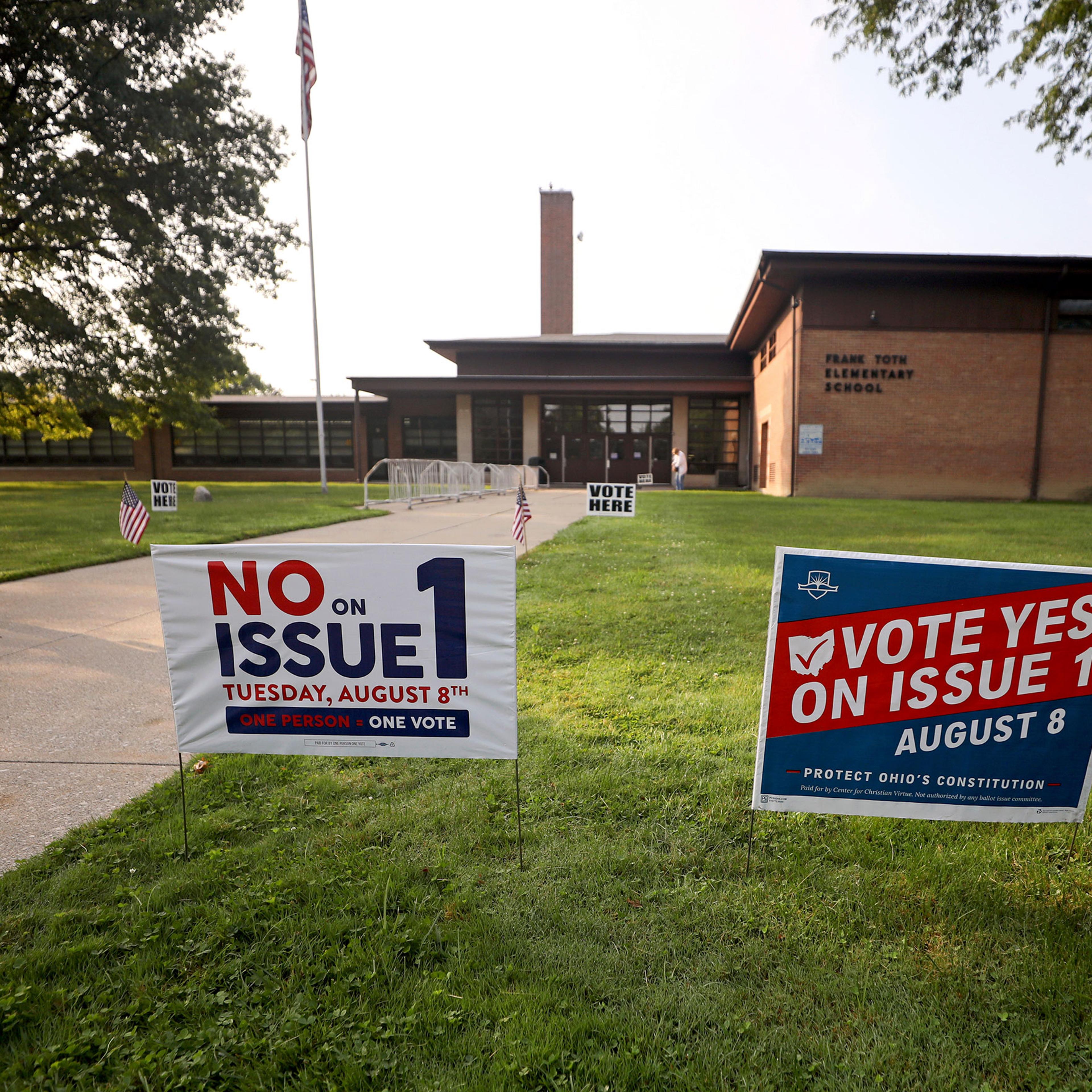 Two opposing signs with slogans posted in the green grass in front of an elementary school where people are going in to vote