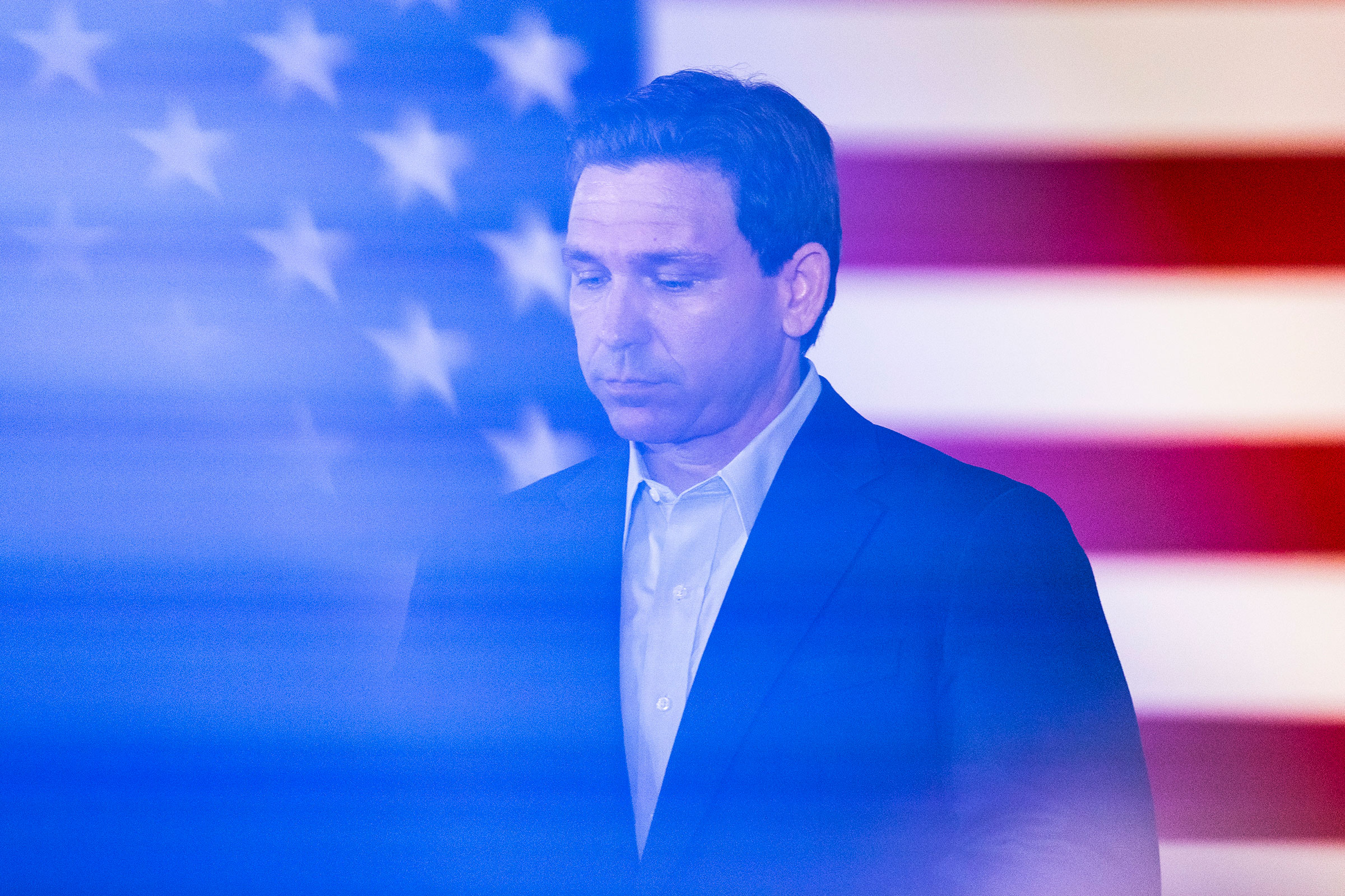 DeSantis with his eyes casting down stands in front of an American flag. There is a blue cast over the image