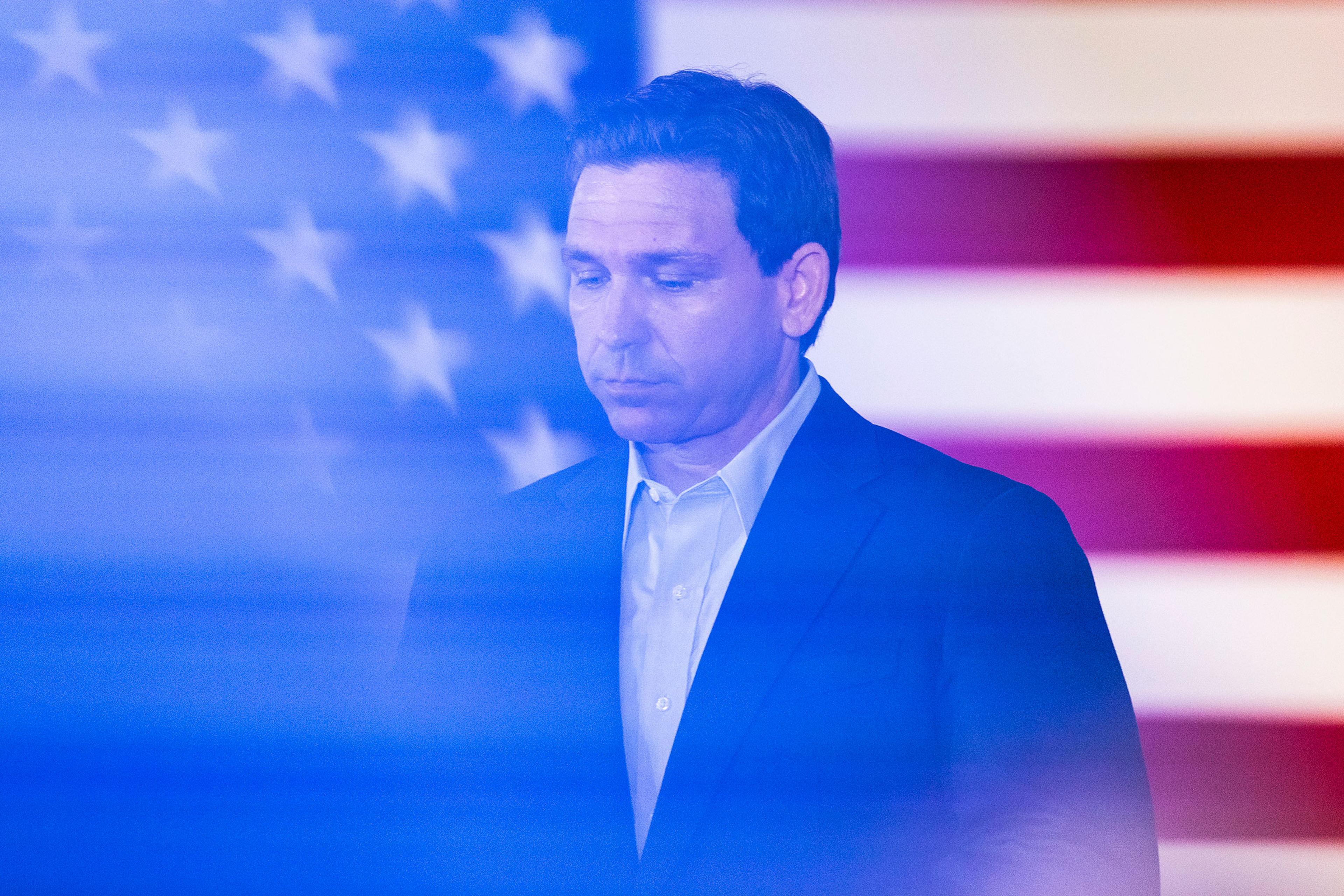 DeSantis with his eyes casting down stands in front of an American flag. There is a blue cast over the image