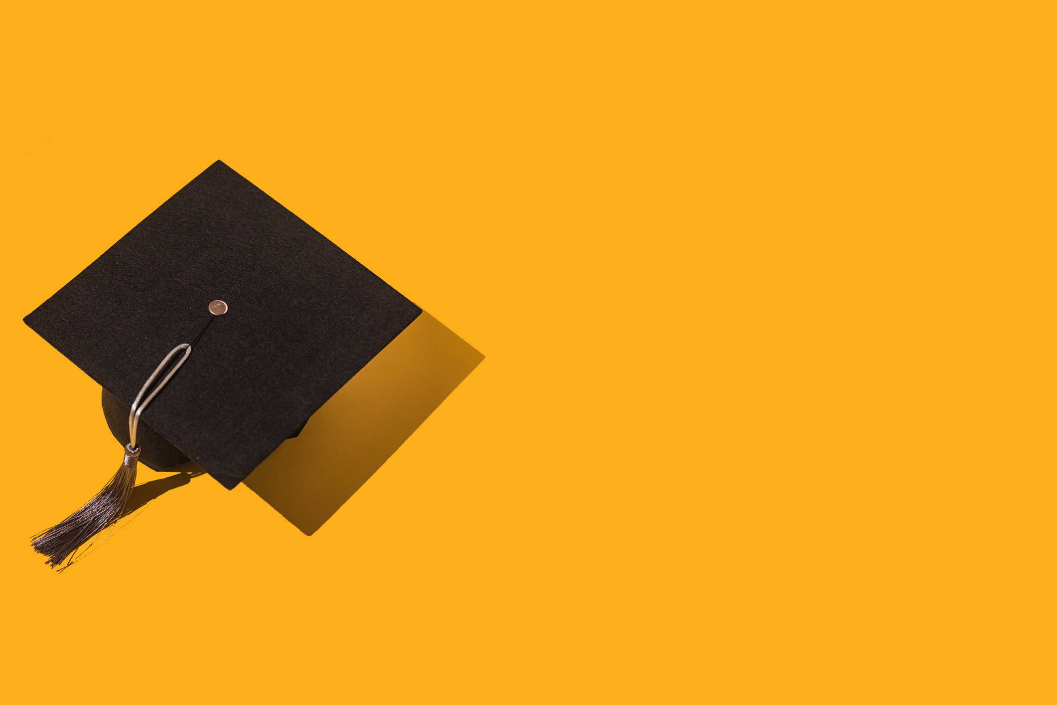 Black graduation cap with gray tassel with hard shadow, on the left side, on yellow background. Graduation, achievement, goal, degree, master, bachelor, university, college and success concept.