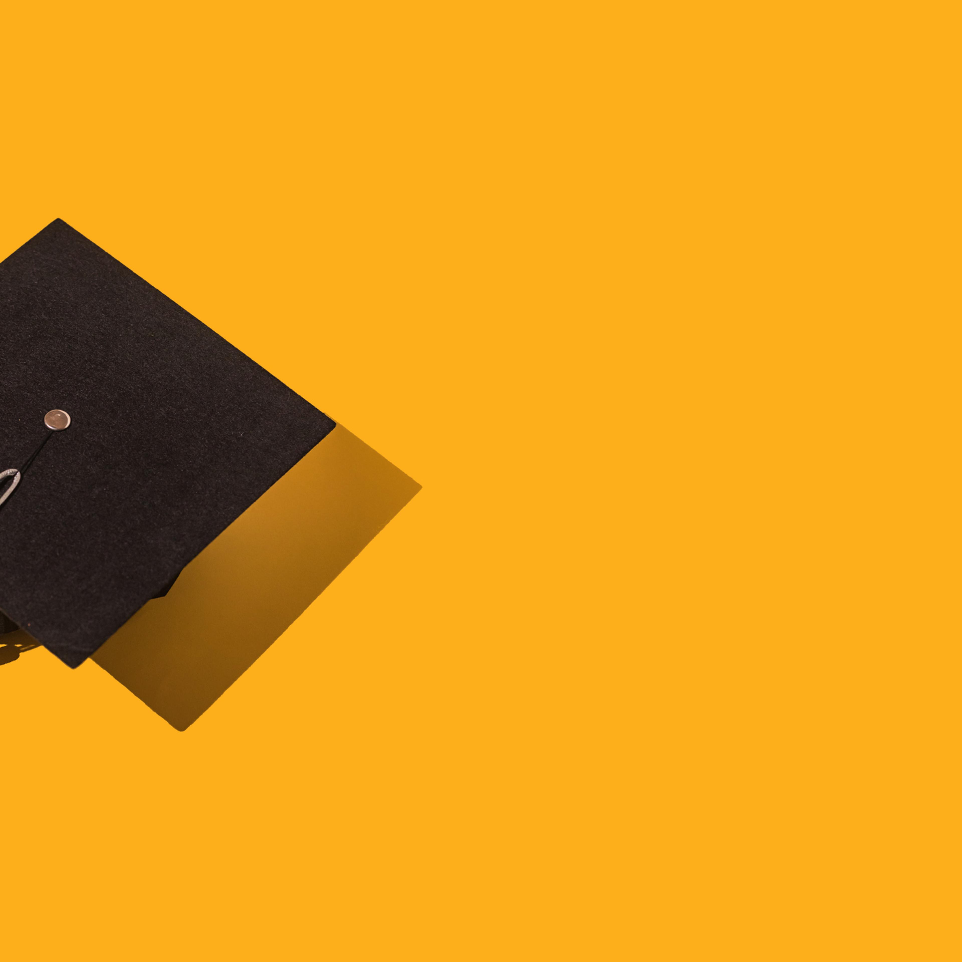 Black graduation cap with gray tassel with hard shadow, on the left side, on yellow background. Graduation, achievement, goal, degree, master, bachelor, university, college and success concept.