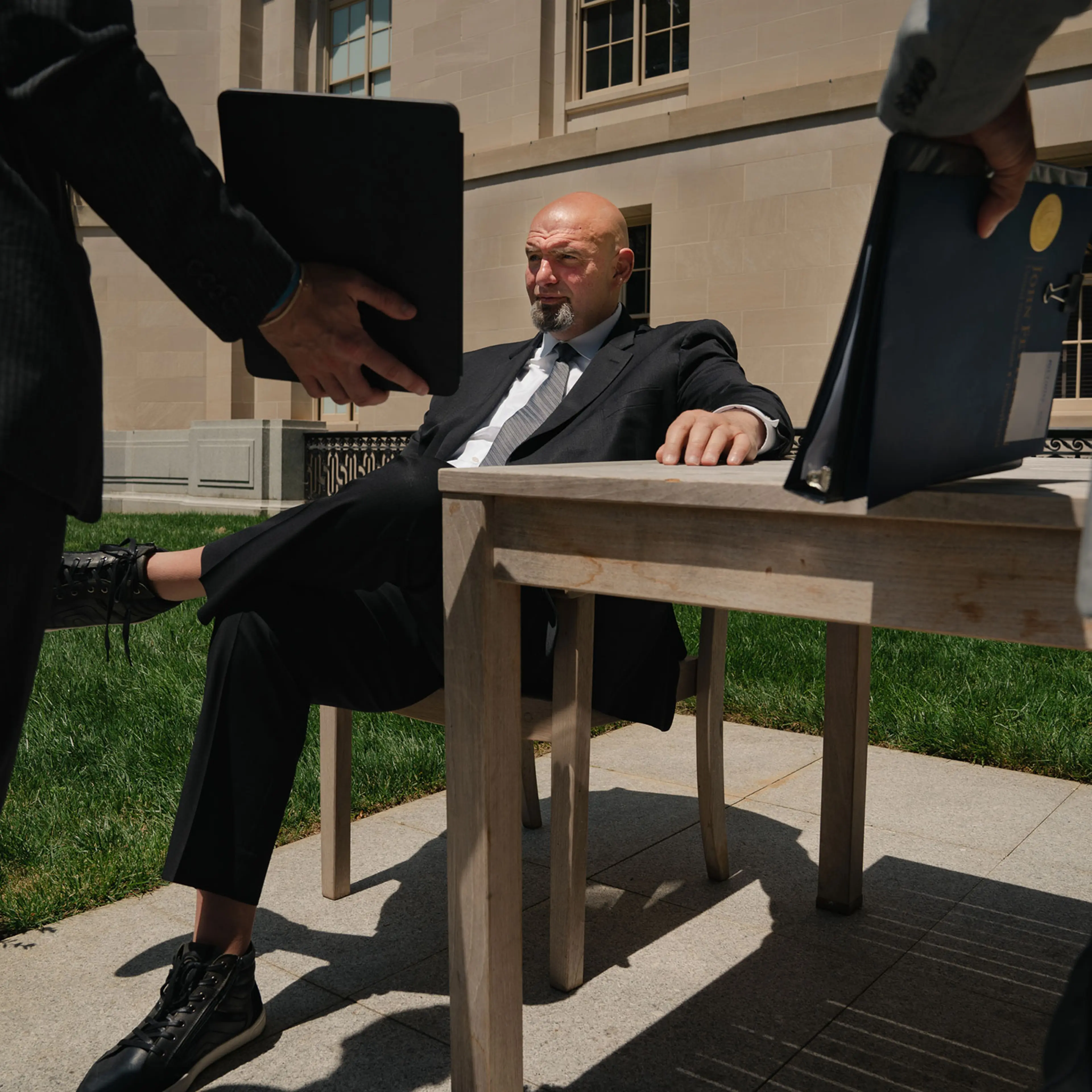 Senator John Fetterman sits in a chair outside in a courtyard in the sun as two other men show him a binder and an ipad, which has a prompter on it so he can have captions of what people around him are saying