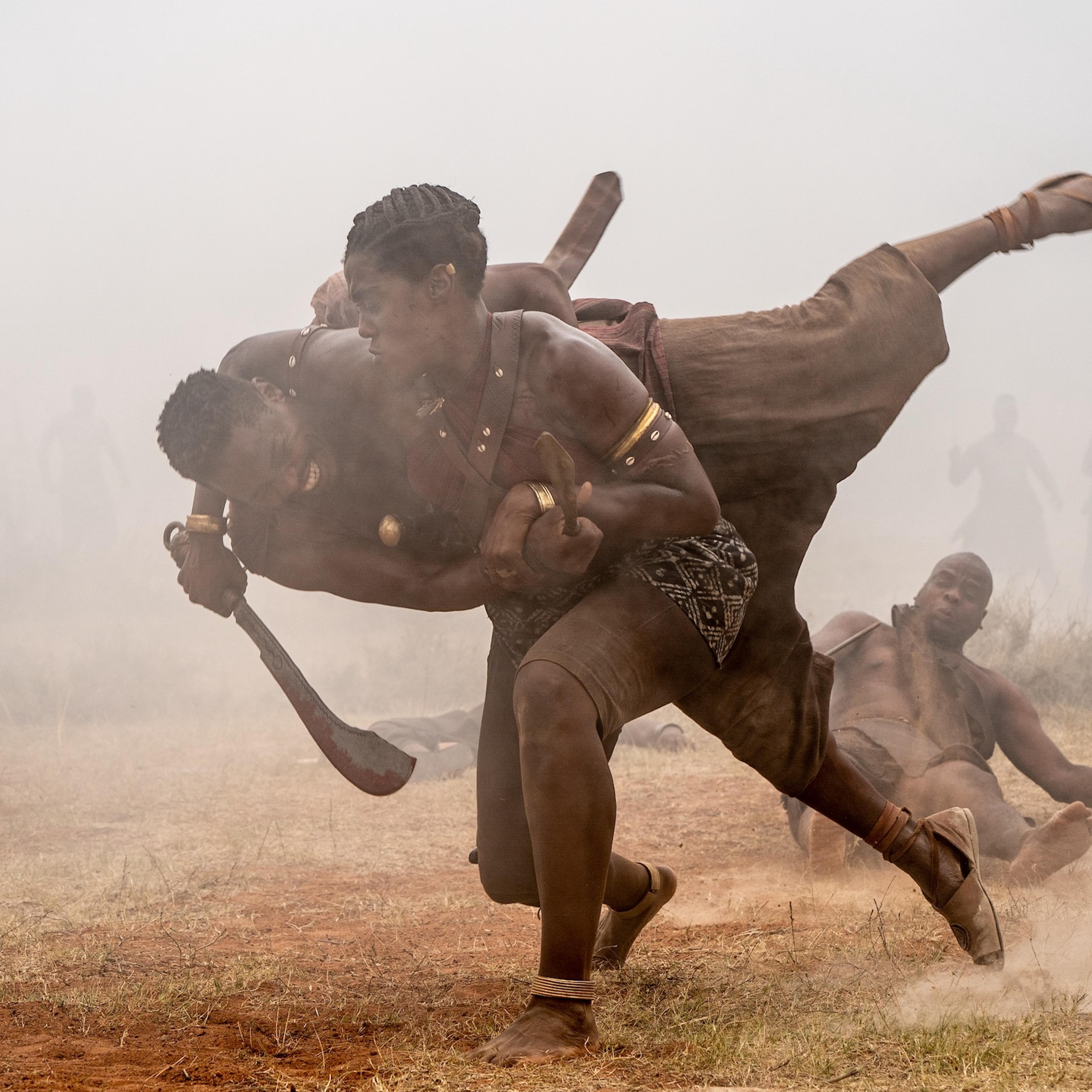 An African woman warrior tackles an African man onto the ground, sword wielded, in battle.