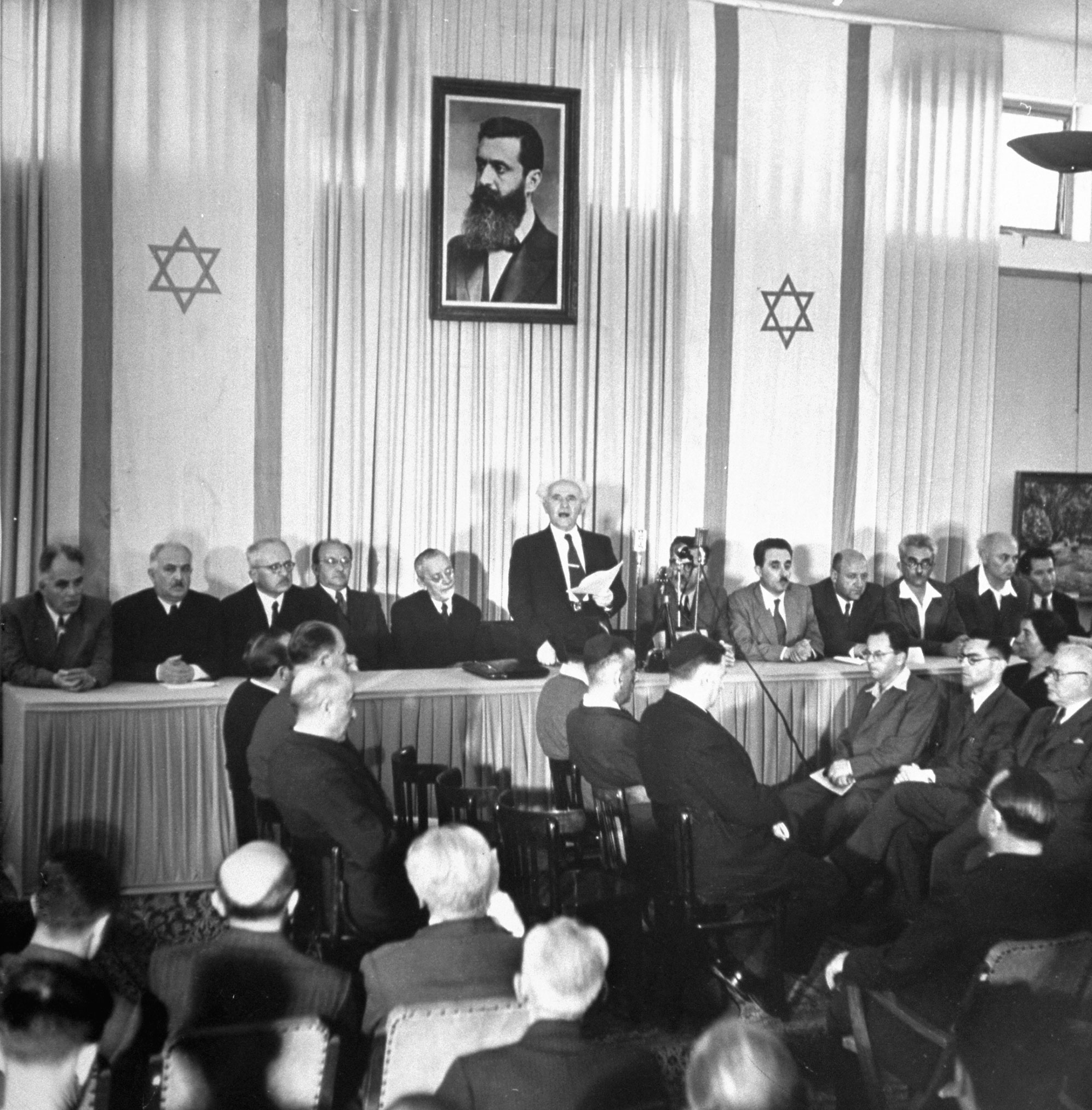 Proclamation of Nationhood is read by Israel's Prime Minister David Ben-Gurion. Around him are members of the provisional government, including Foreign Minister Moshe Shertok (third from right). Labor Minister Moshe Ben Tov (extreme right) wears sport sh