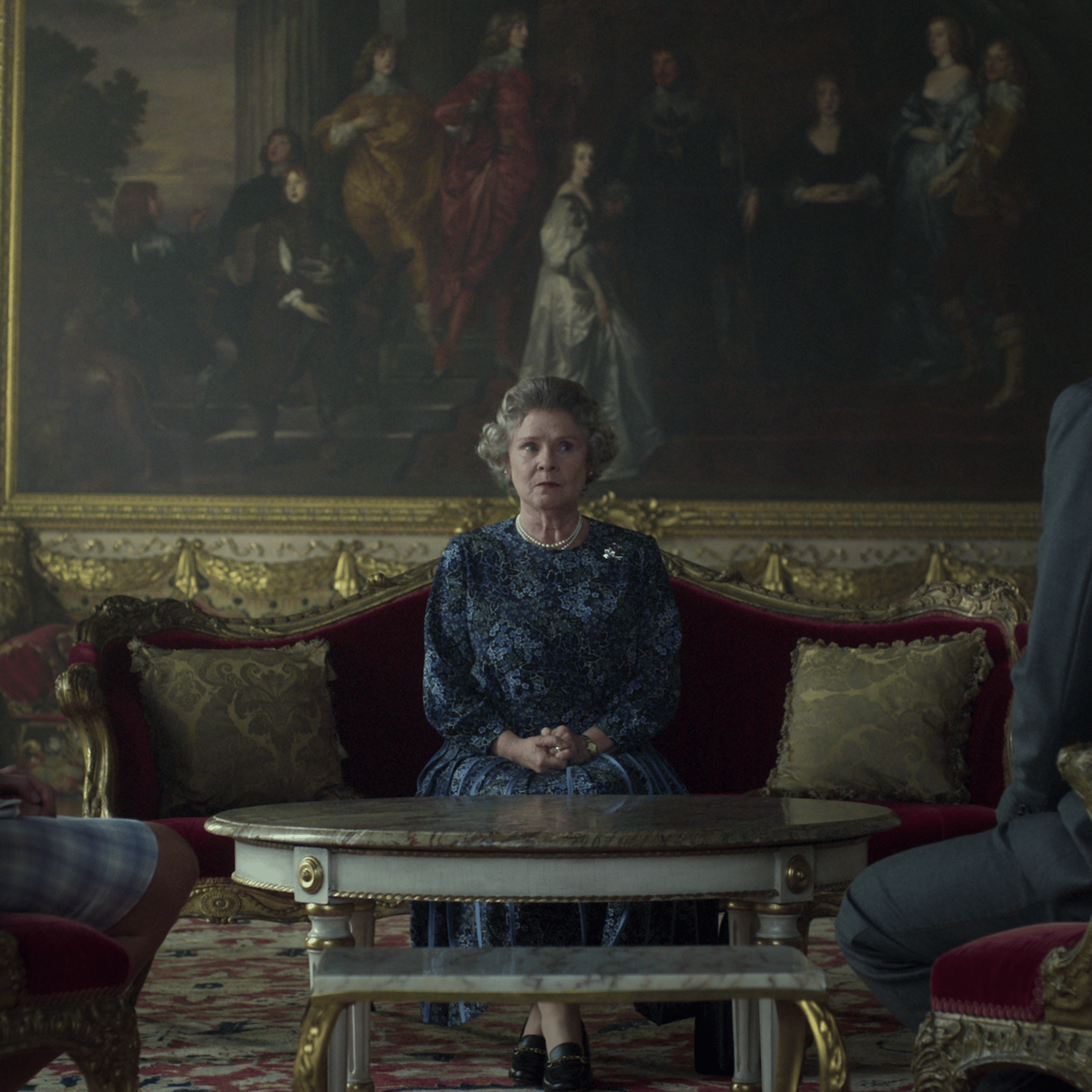 Queen Elizabeth II sits on a red velvet couch with an advisor on either side.