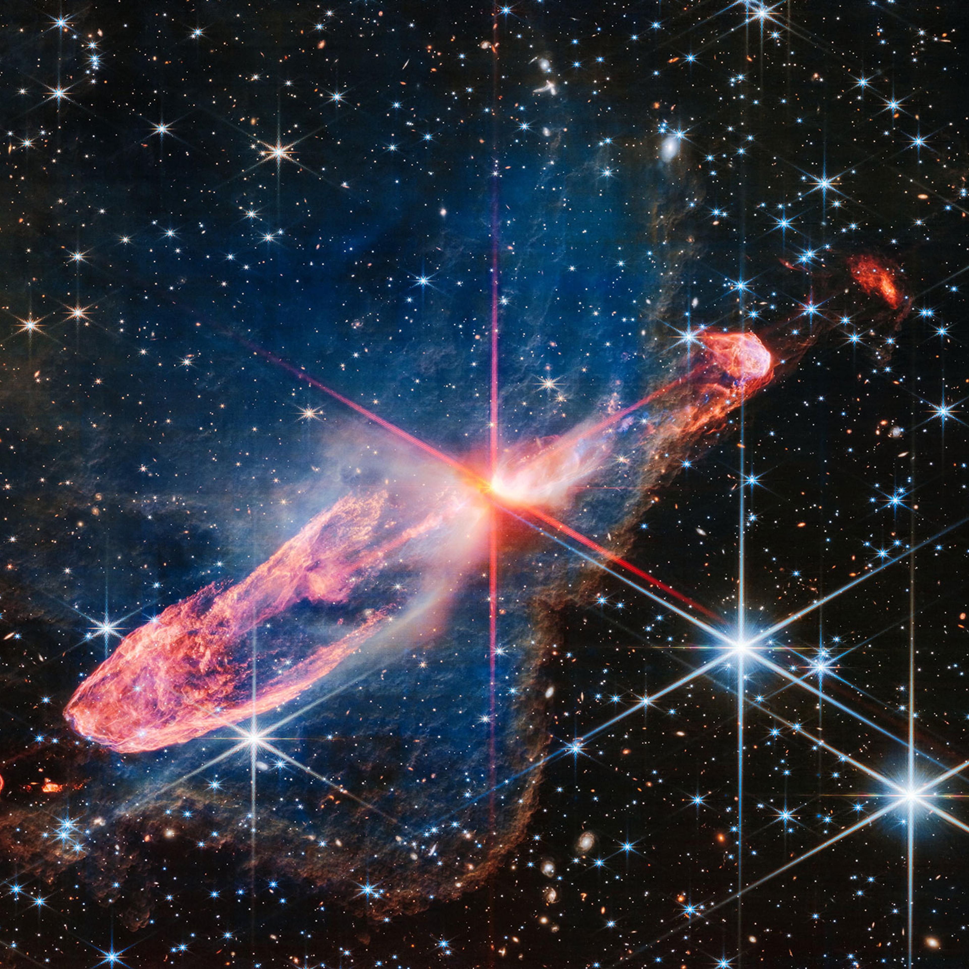 At the center of the image is a horizontal orange cloud known as Herbig-Haro 46/47 that is uneven with rounded ends, and tilted from bottom left to top right. In the middle of this cloud is a yellow-white blob with 8 reddish pink diffraction spikes pierc