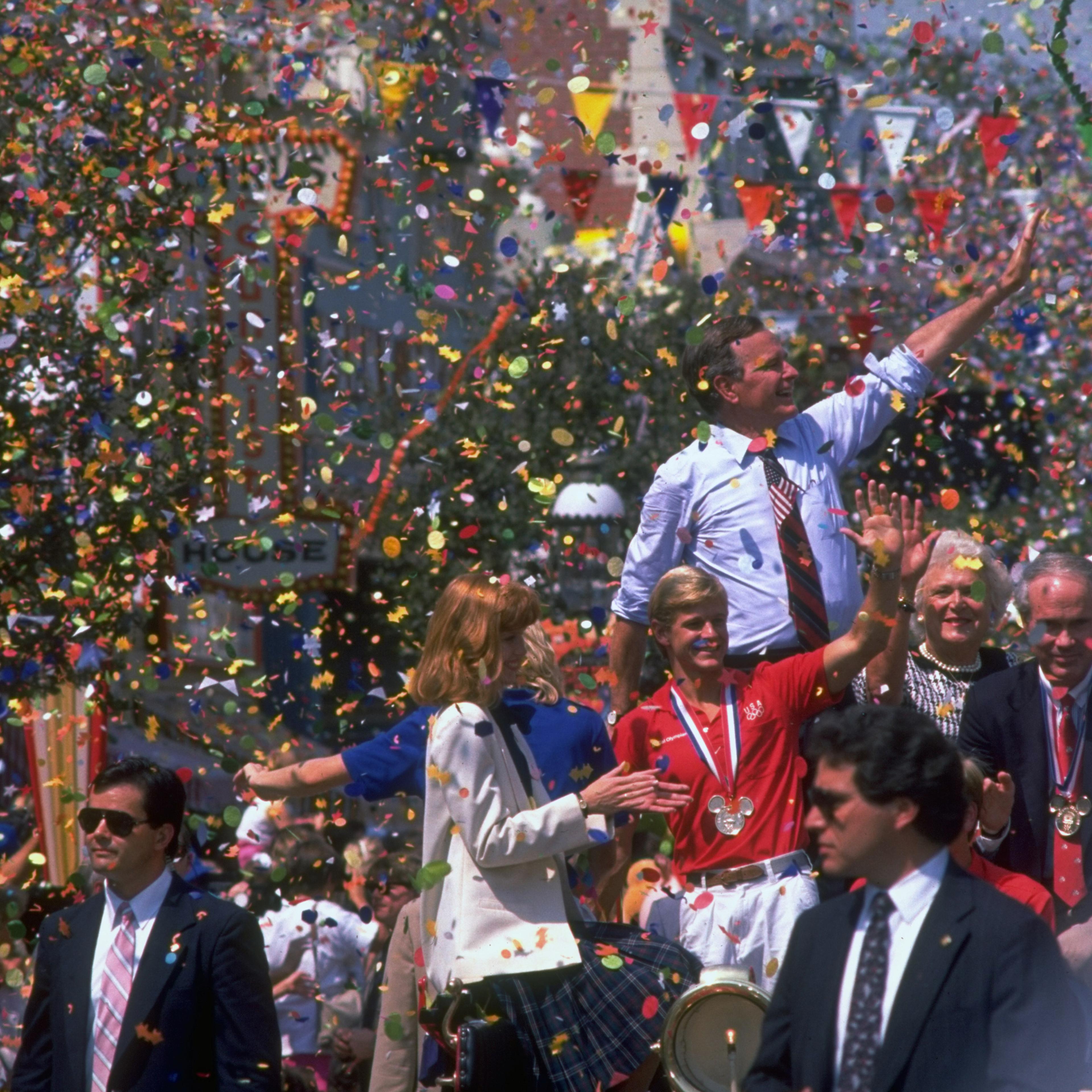 Republican presidential candidate George H.W. Bush waving above a crowd amid shower of parade confetti, with wife Barbara by his side at a send-off gala for the US Olympic team.