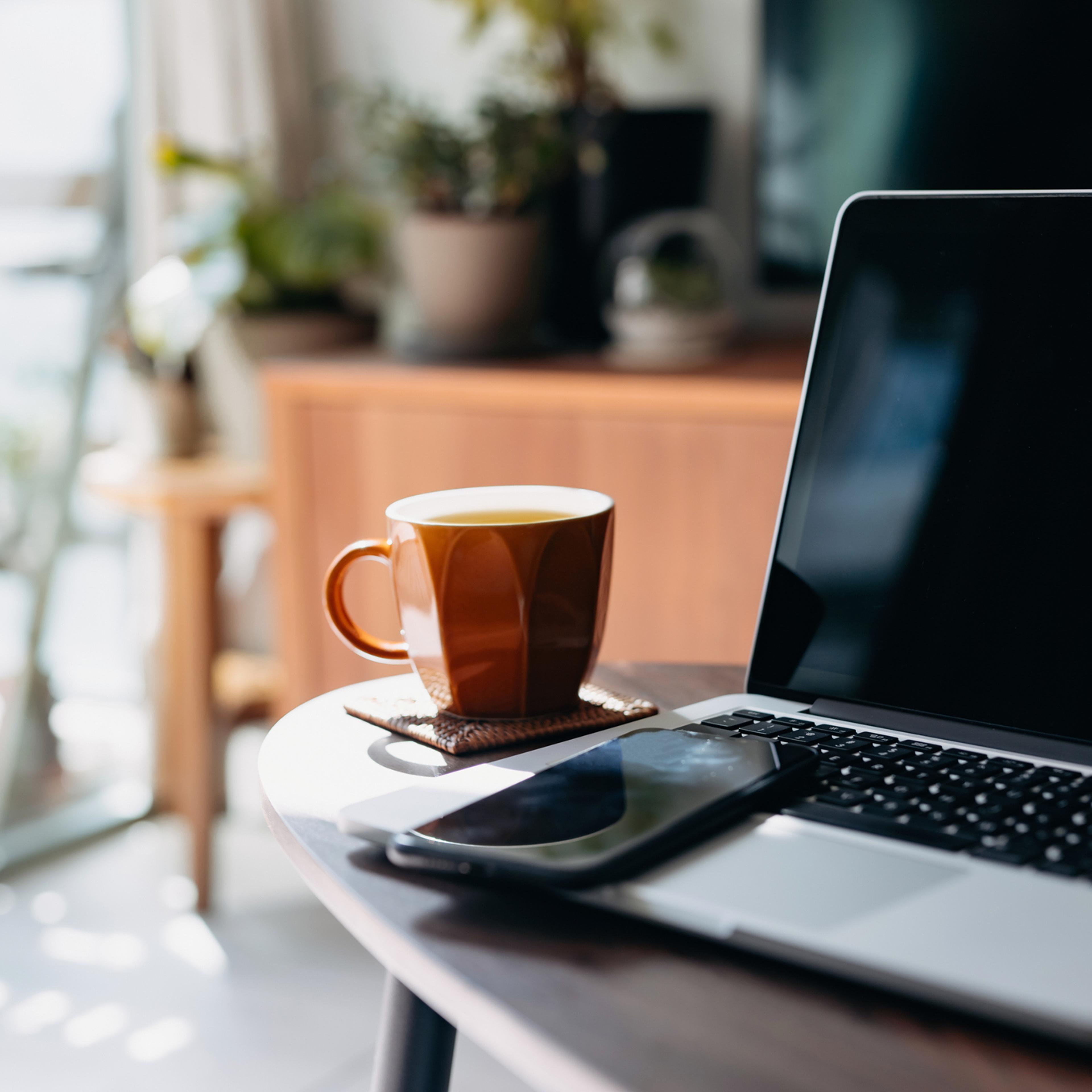 Cropped shot of wooden coffee table with laptop, smartphone and a cup of tea in the living room at home by the window against beautiful sunlight