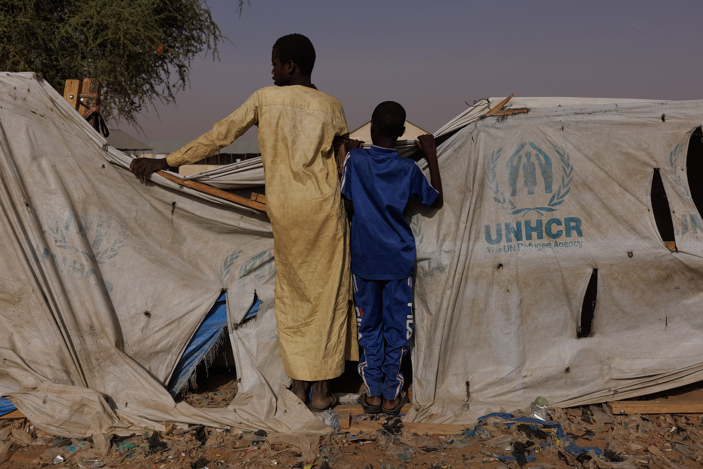 Two boys peer over a UNHCR aid tent as newly arrived refugees gather at a relocation camp near the border in Adre, Chad, on April 19, 2024.