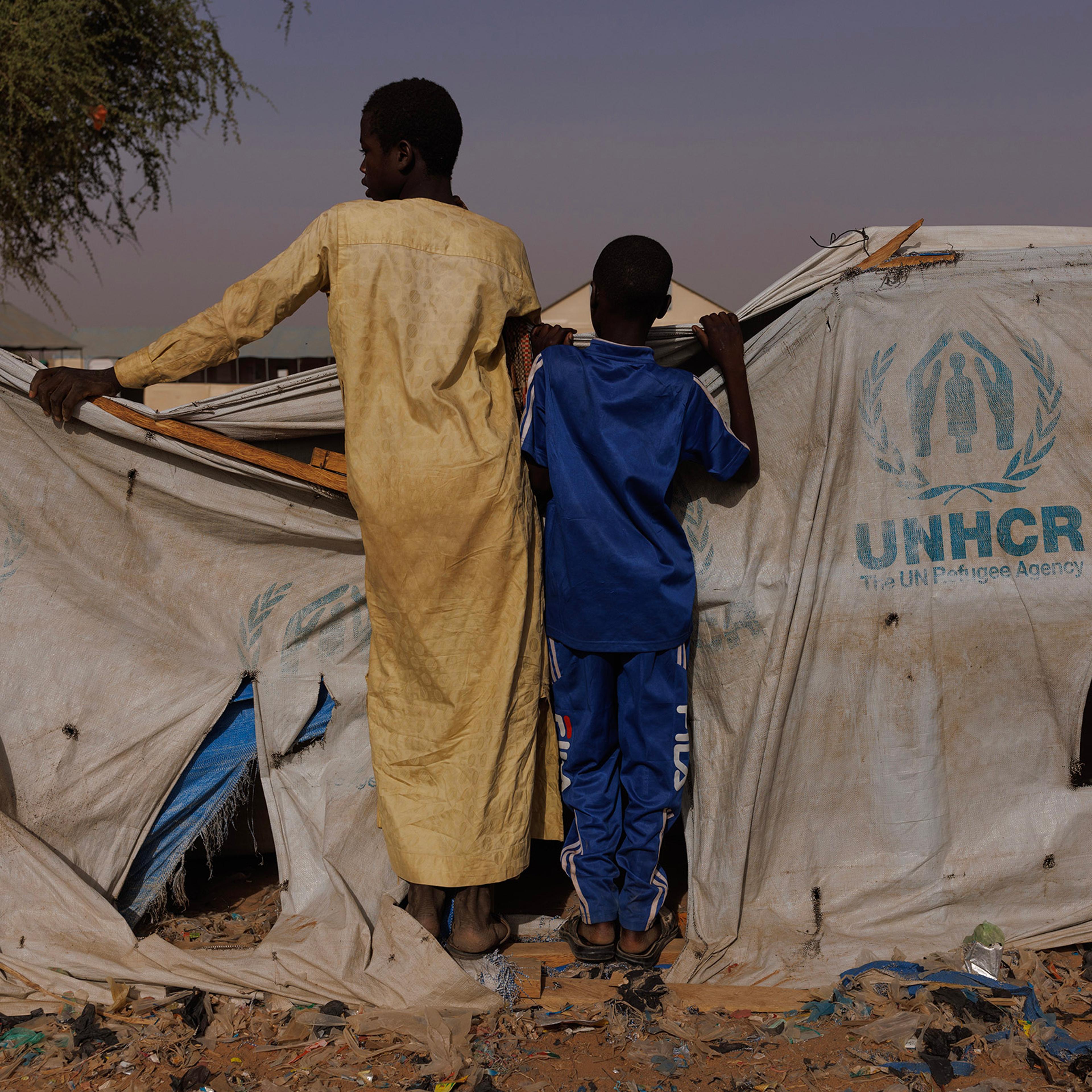 Two boys peer over a UNHCR aid tent as newly arrived refugees gather at a relocation camp near the border in Adre, Chad, on April 19, 2024.