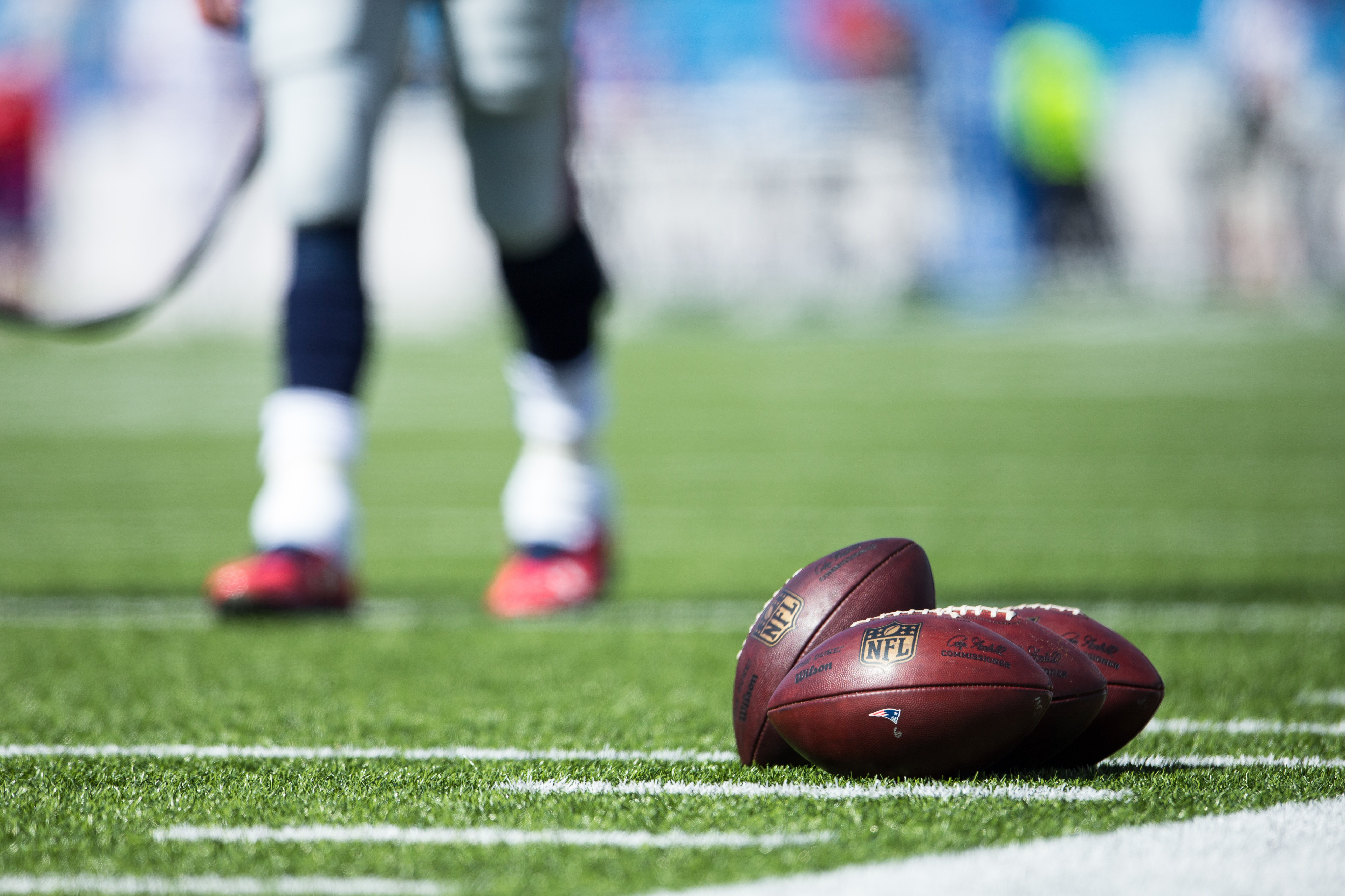 ORCHARD PARK, NY - SEPTEMBER 20:  Wilson NFL Footballs bearing the New England Patriots logo set on the turf as Tom Brady #12 warms up in the background before the game against the Buffalo Bills on September 20, 2015 at Ralph Wilson Stadium in Orchard Pa