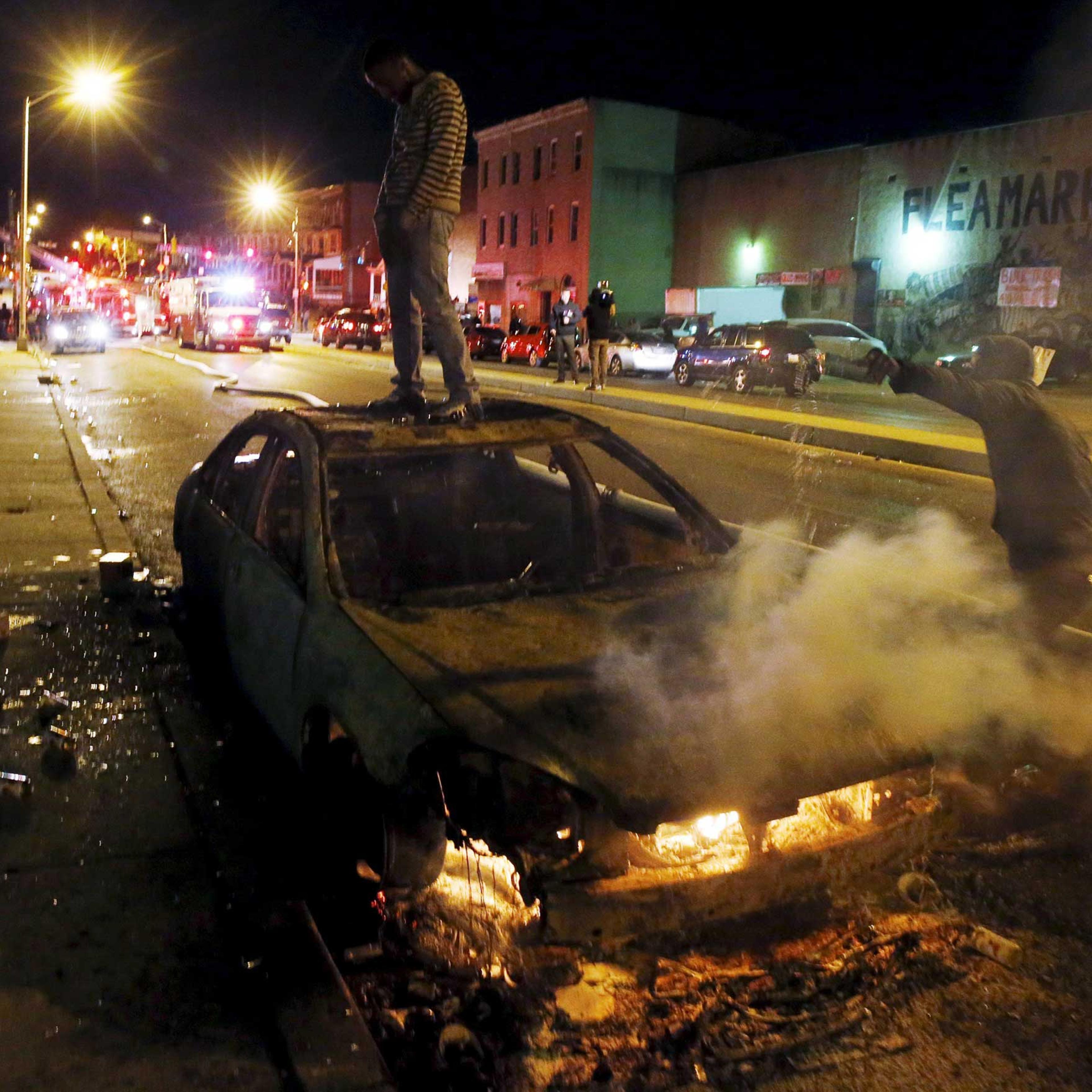 A rioter stands atop a burning car as another man pours fuel onto the fire while Baltimore firefighters behind them fight fires in mutliple burning buildings set ablaze by rioters during clashes in Baltimore