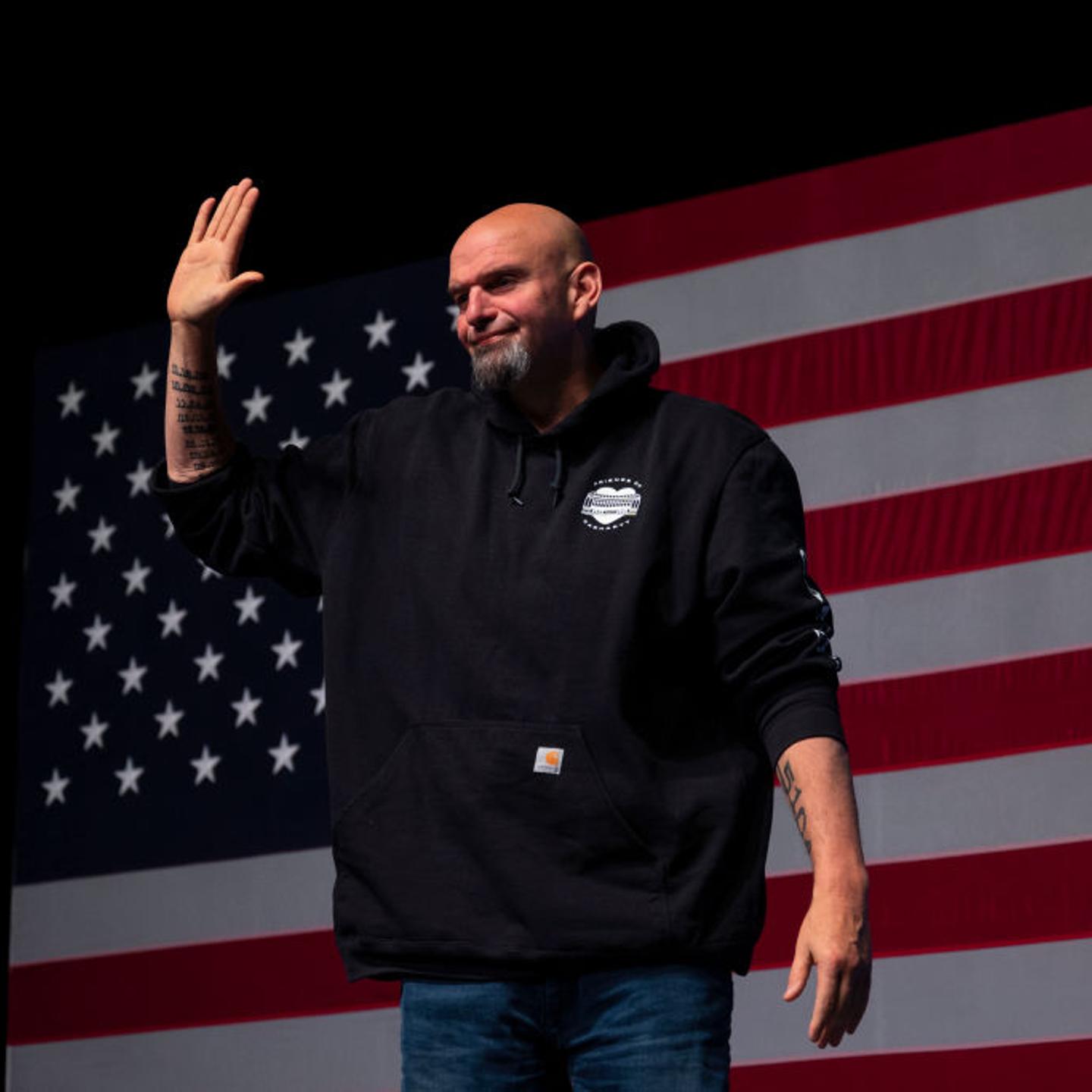 John Fetterman waves his hand as he stands behind an American flag.