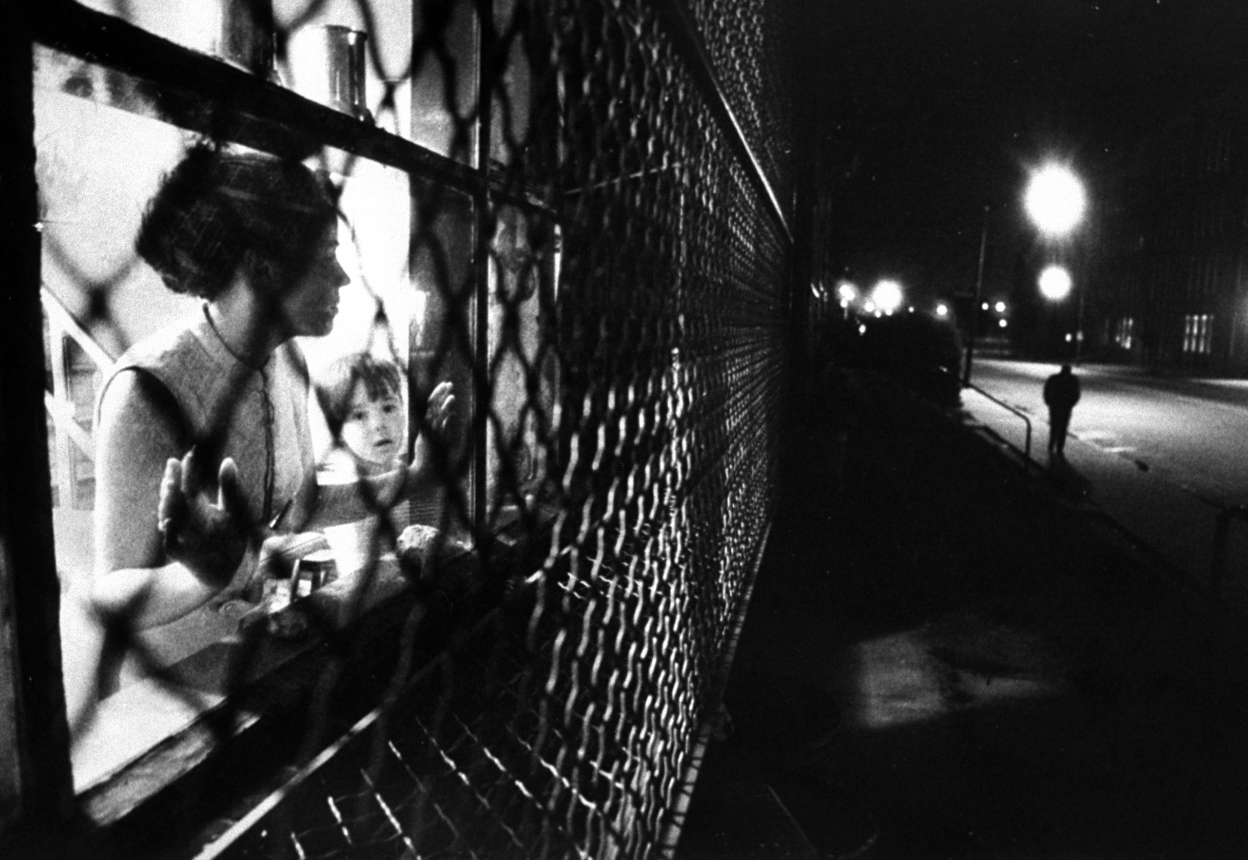 In a Baltimore ghetto, Mrs. Judy Brookhouser watches from the caged window of her apartment in "The Compound" —the guarded complex operated by Johns Hopkins Hospital for families of staff doctors.