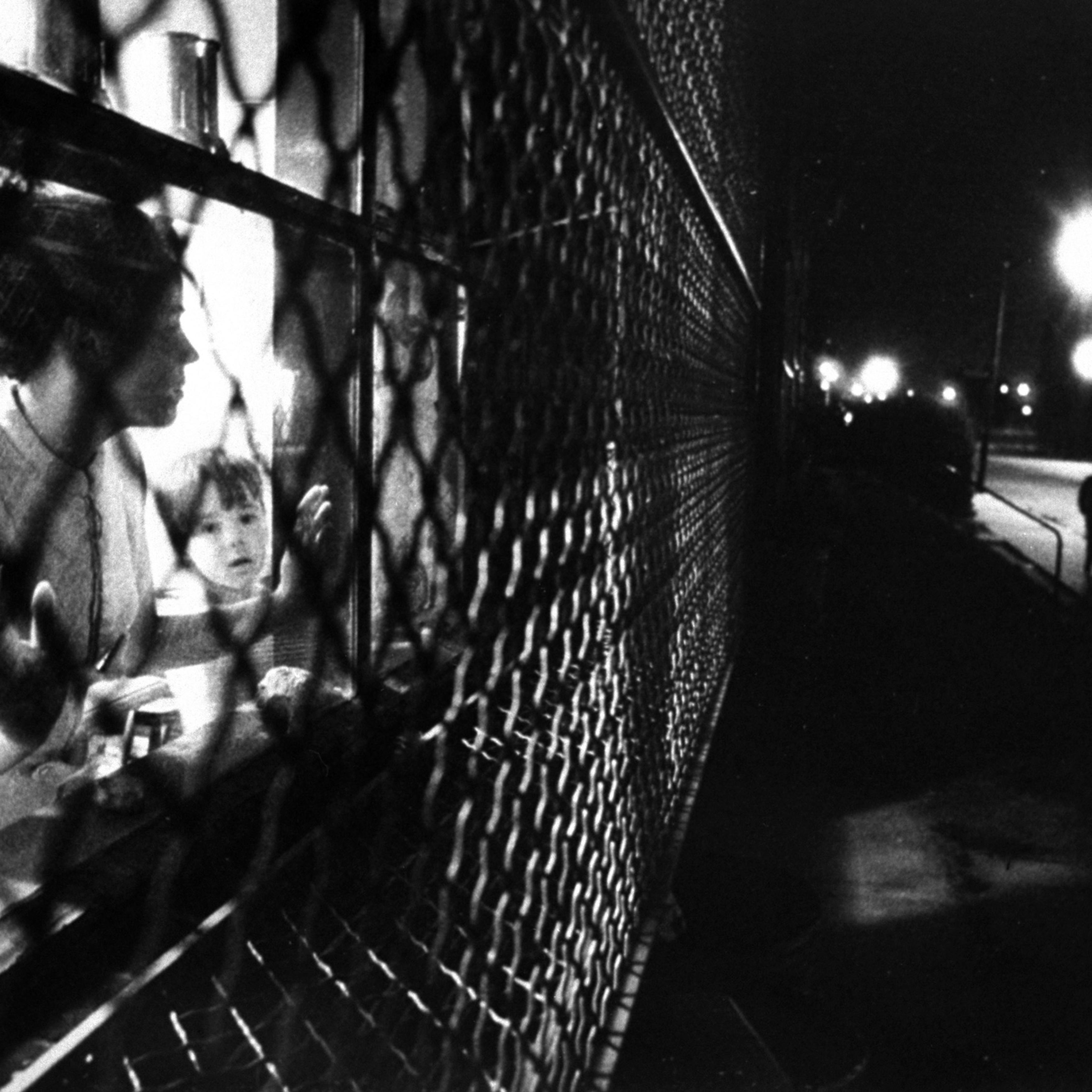 In a Baltimore ghetto, Mrs. Judy Brookhouser watches from the caged window of her apartment in "The Compound" —the guarded complex operated by Johns Hopkins Hospital for families of staff doctors.