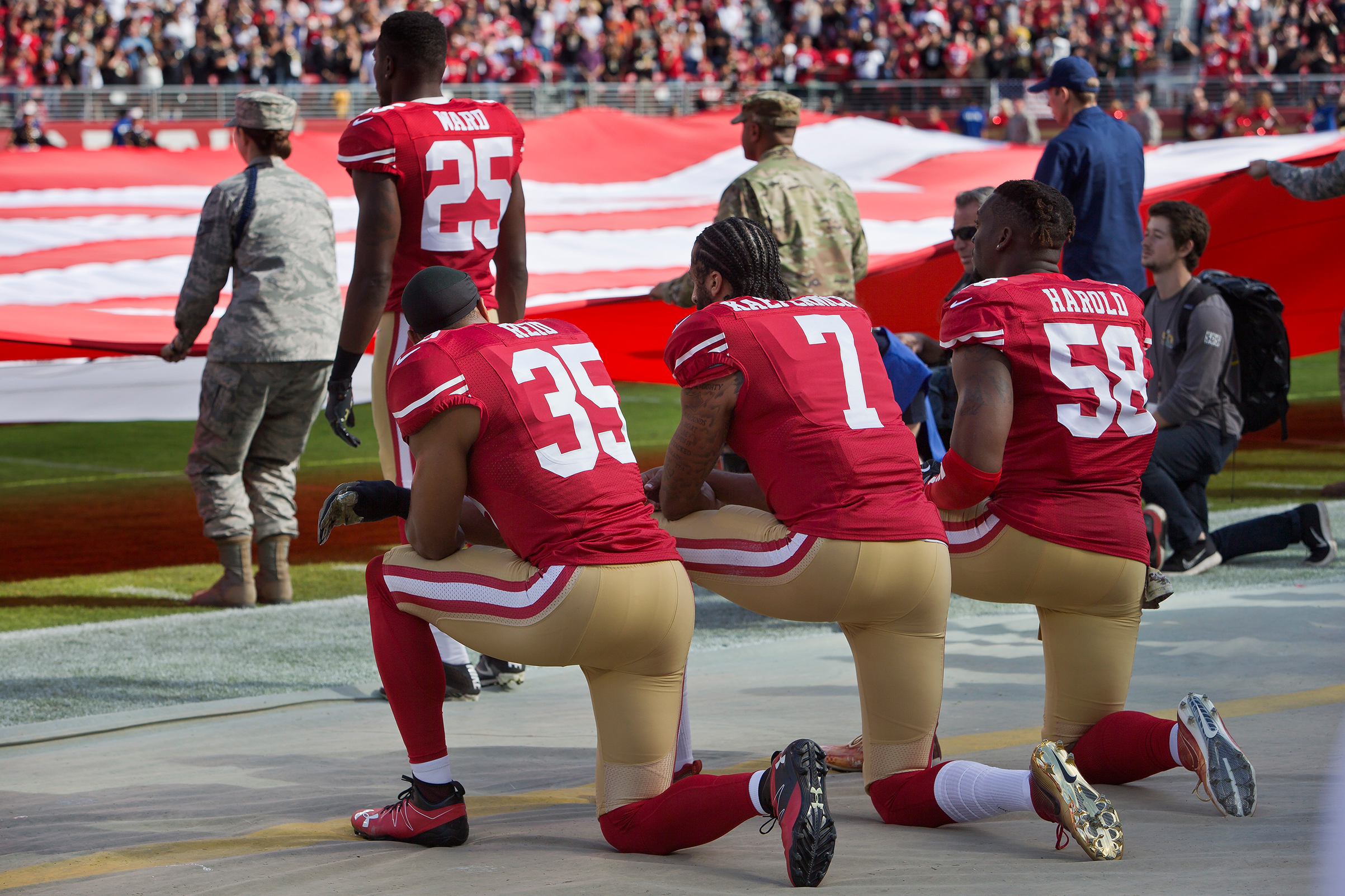 Quarterback Colin Kaepernick #7, safety Eric Reid #35, and linebacker Eli Harold #58 of the San Francisco 49ers kneel before a game against the New Orleans Saints with the U.S. flag unfurled in honor of the armed services on Nov. 6 2016 at Levi's Stadium