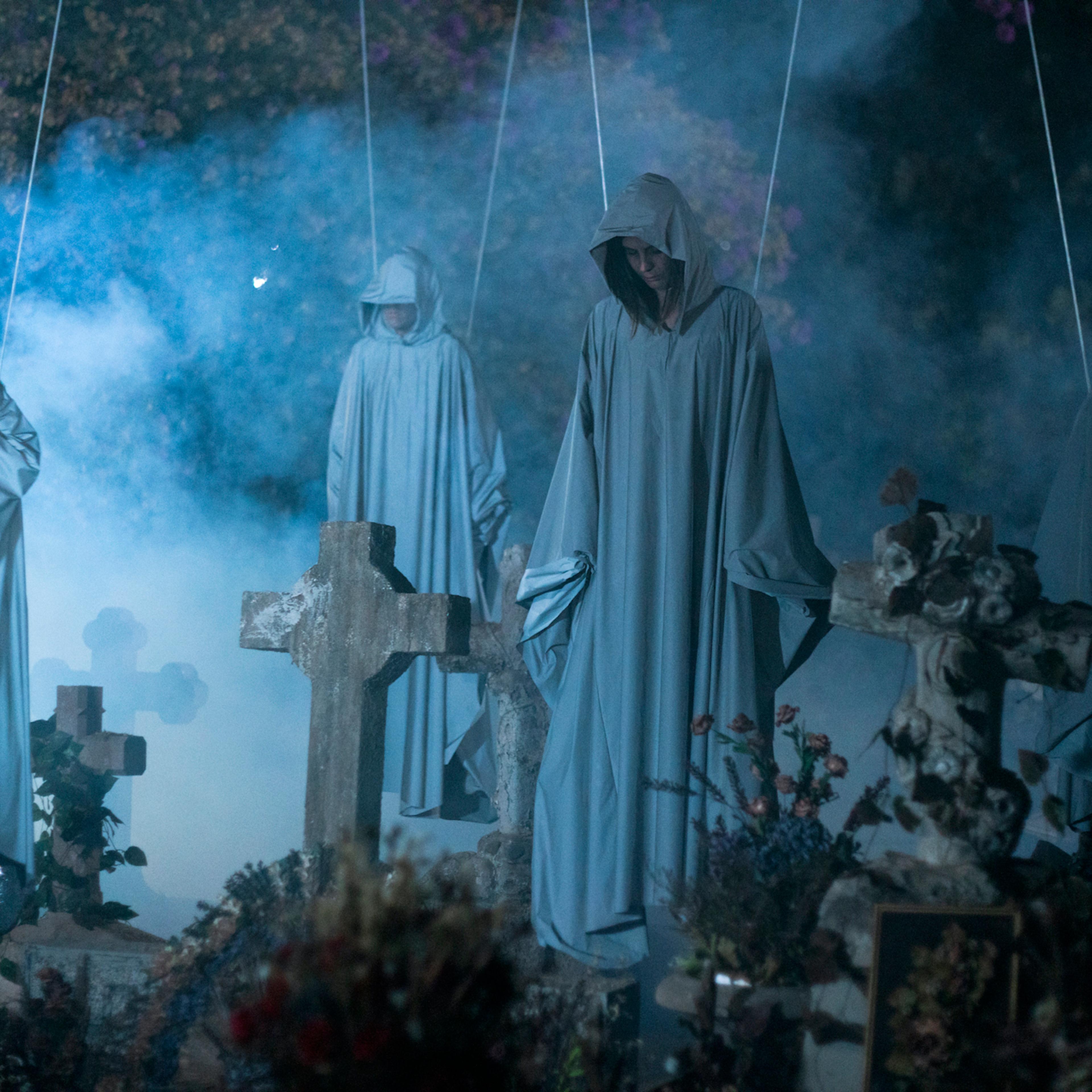 Four people in gray robes hang, suspended from ropes, above a cemetery.