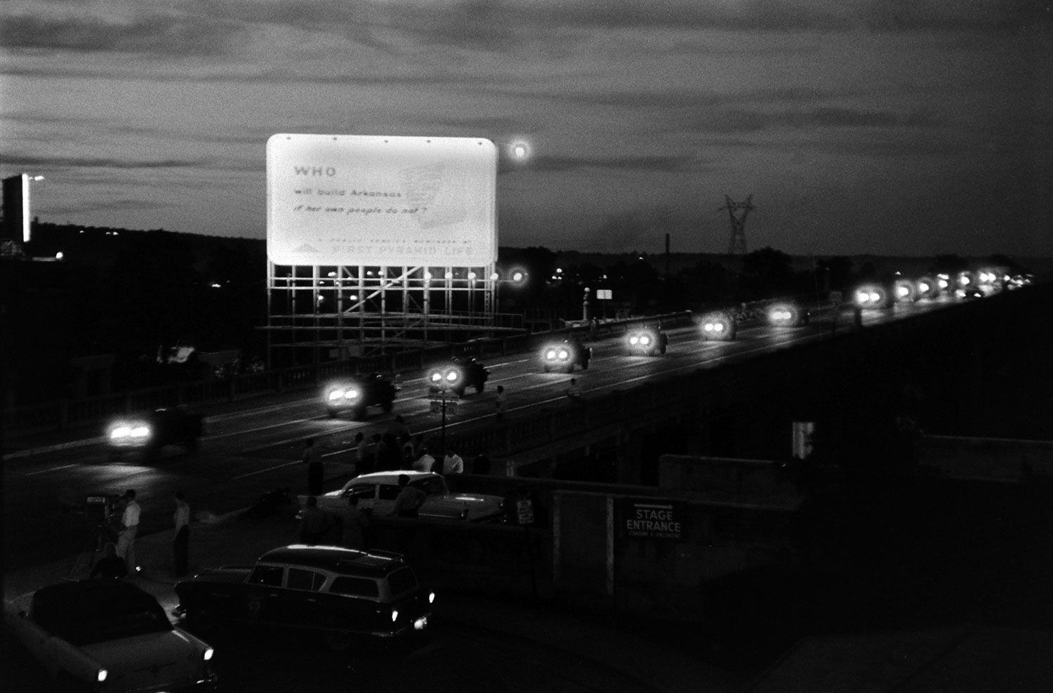 A convoy of Jeeps from the 101st Airborne heads to Little Rock. The billboard they pass reads, "Who will build Arkansas if her own people do not?"