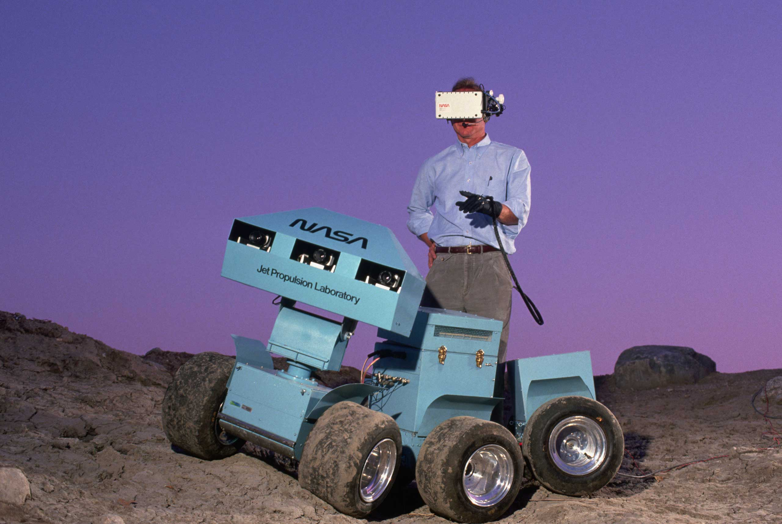 Andrew Mishkin wearing a 3-D virtual display helmet that is connected to a six-wheeled roving vehicle. The rover was meant to explore the surface of Mars and send back information.