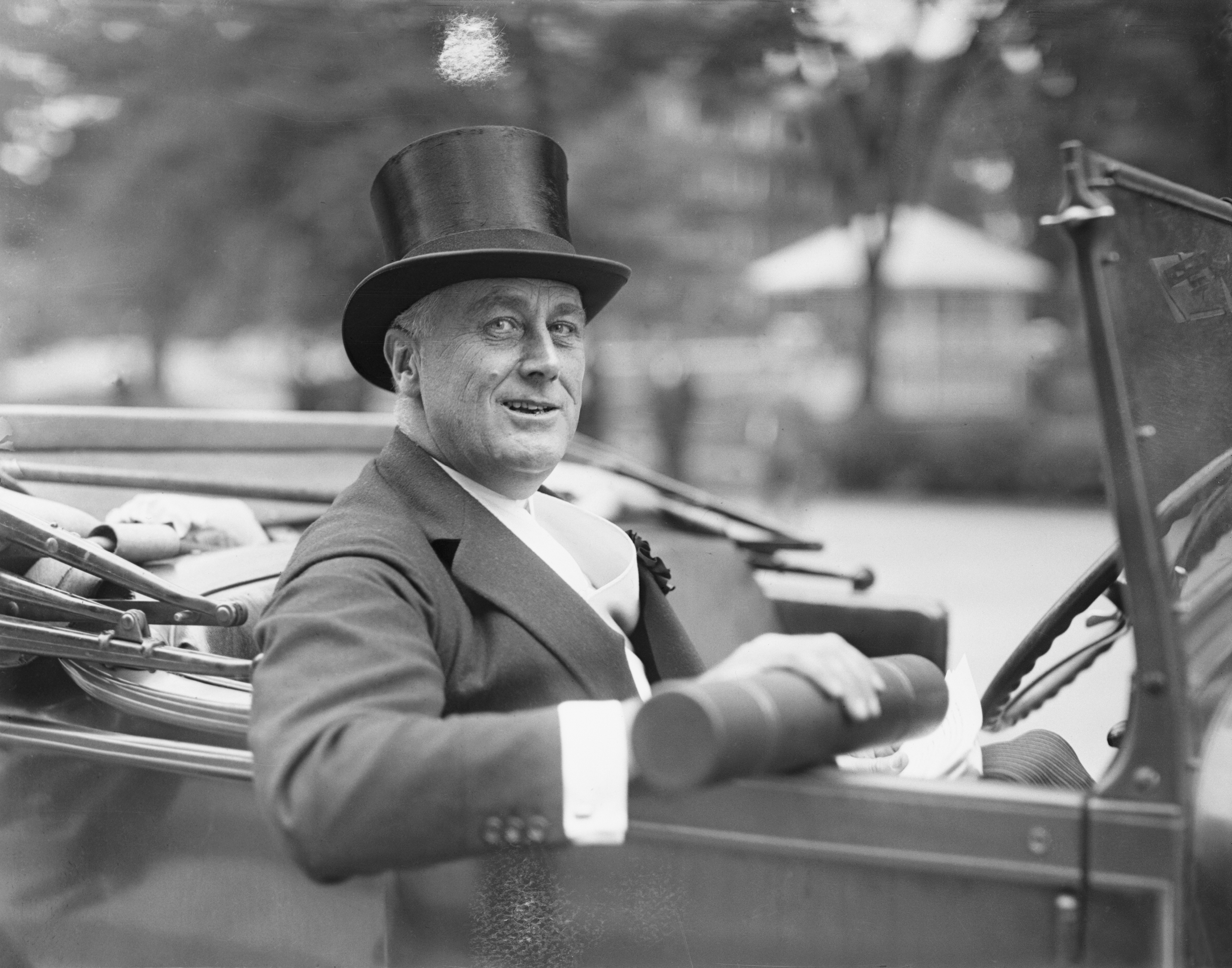 New York Governor Franklin Delano Roosevelt sits in a car, smiling with his top hat on.