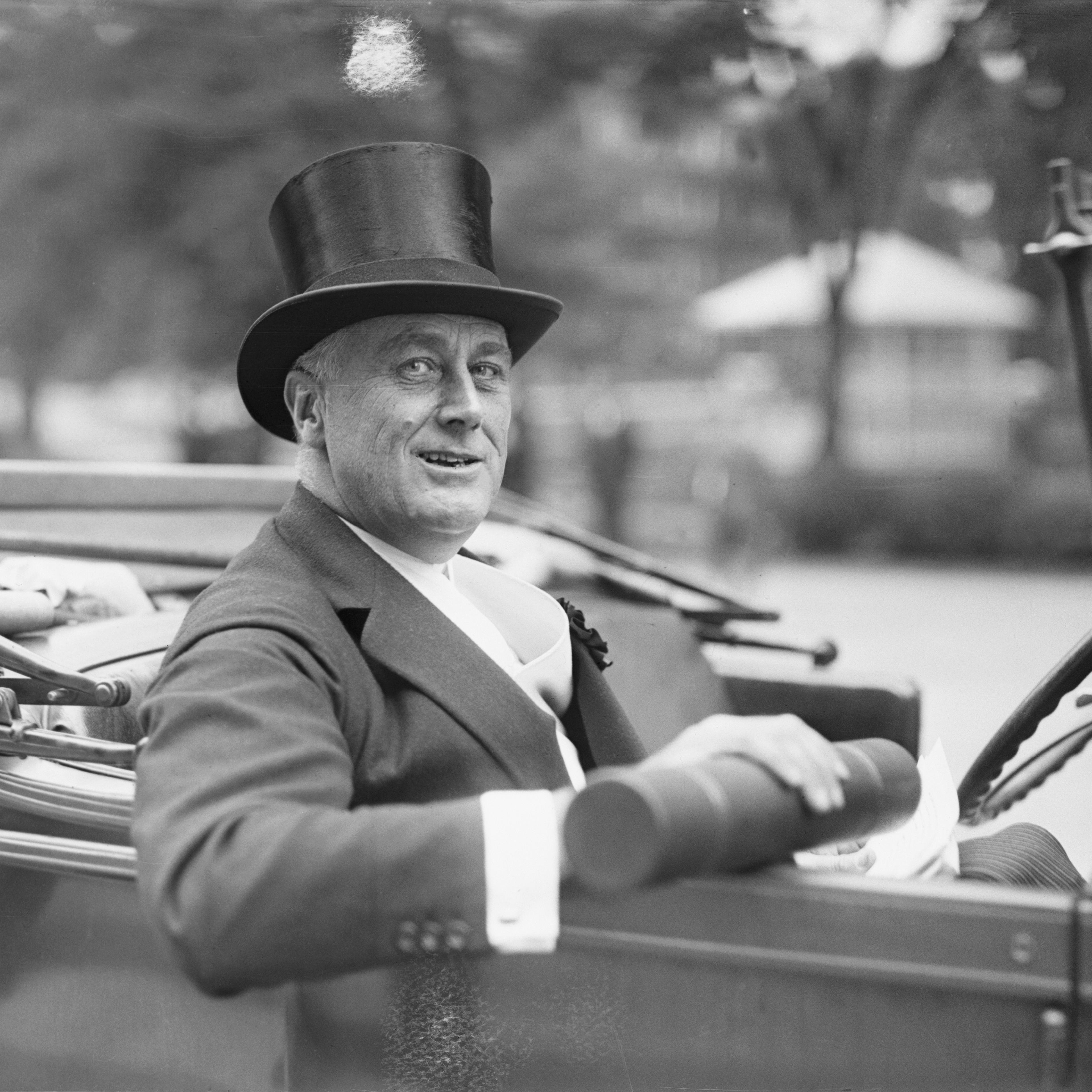 New York Governor Franklin Delano Roosevelt sits in a car, smiling with his top hat on.