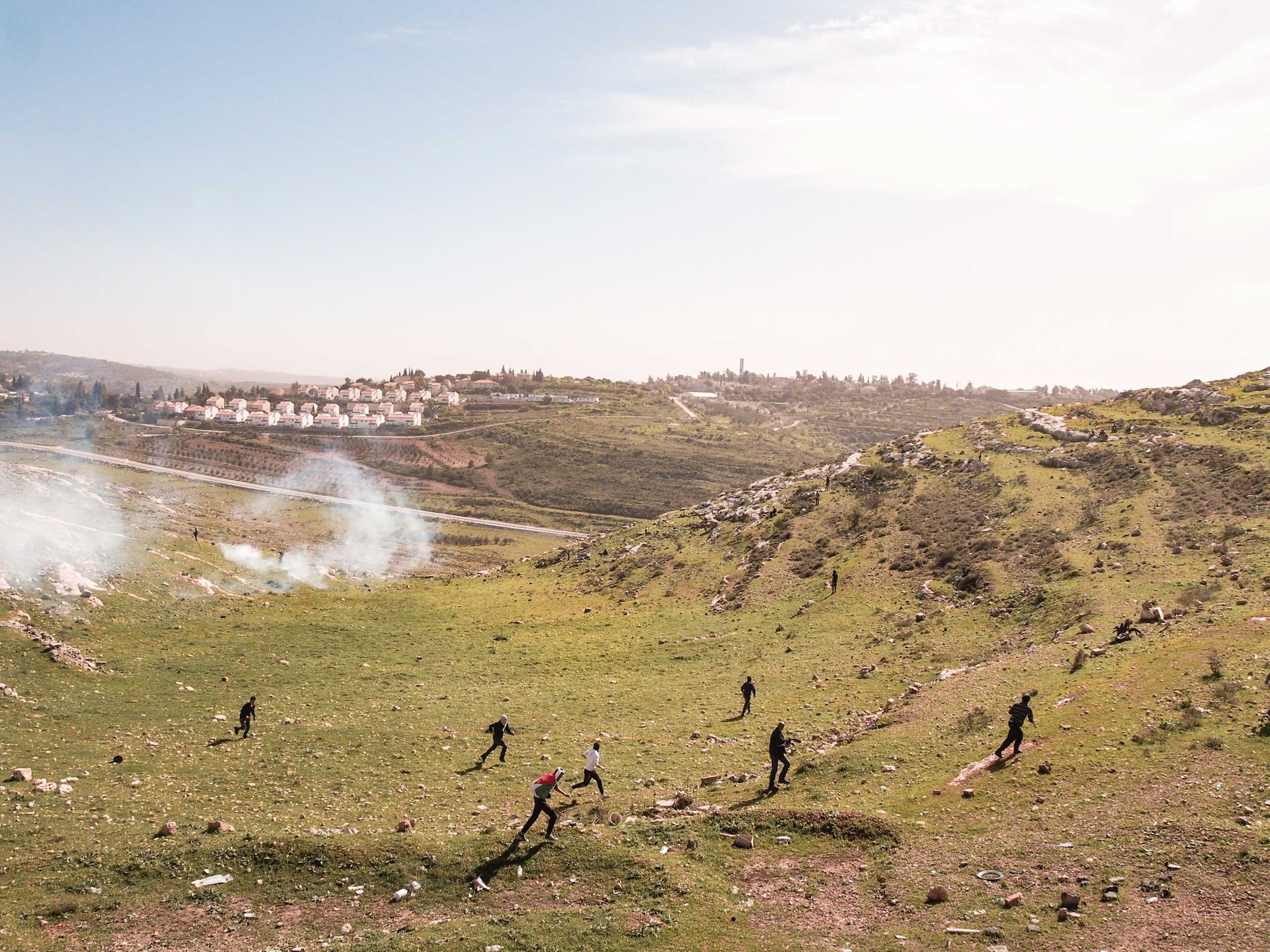 West Bank, Nabi Saleh. 2013. Protestors run away as Israeli soldiers fire tear gas at them. The Israeli settlement of Halimish is in the background.Residents of Nabi Saleh have been protesting the Israeli Occupation every Friday after midday prayers sinc