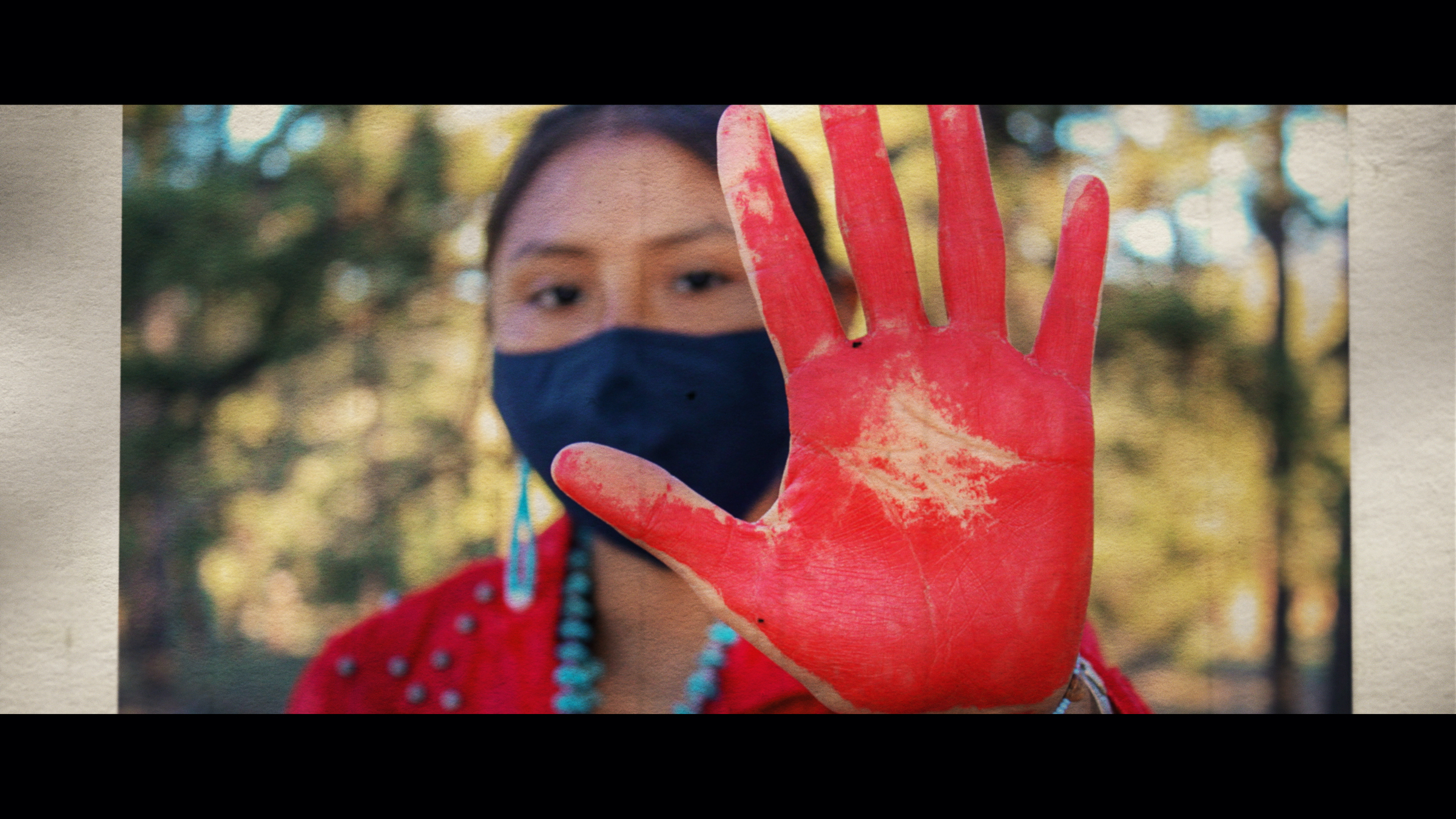 A young Native American woman holds out her open palm, painted red, toward the camera in the foreground.