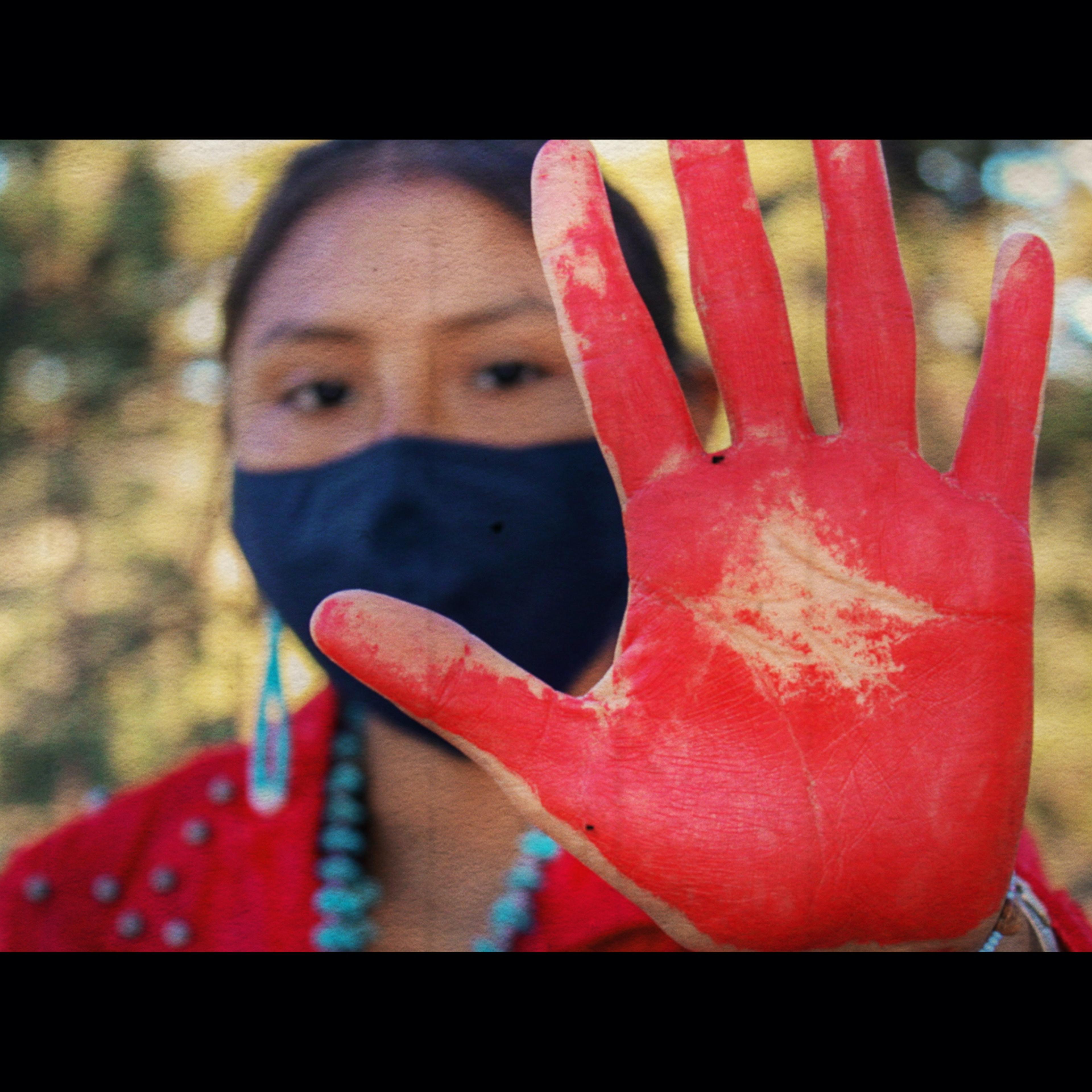A young Native American woman holds out her open palm, painted red, toward the camera in the foreground.