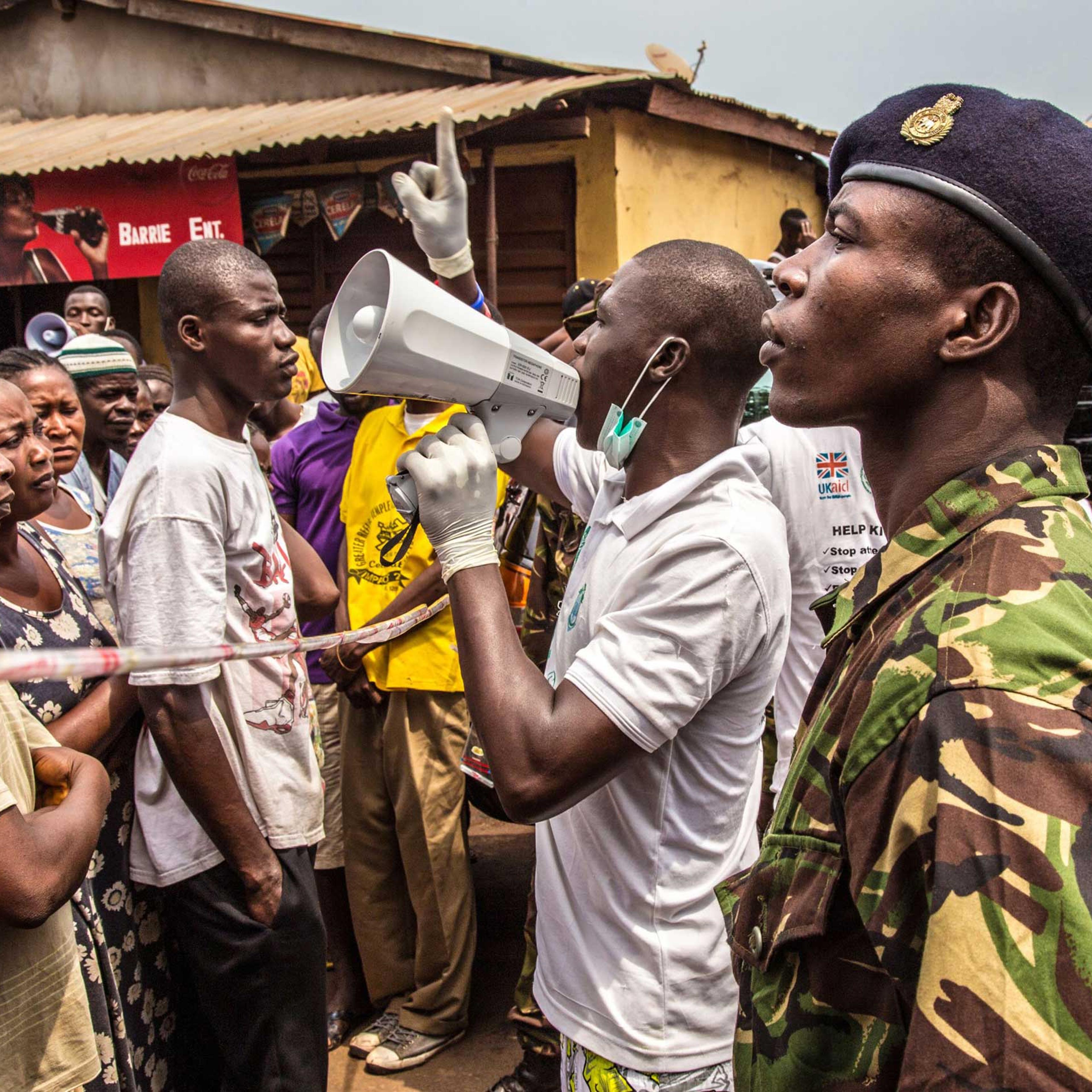People stand in line for food to be distributed to them as a health worker makes an announcement in Freetown, Sierra Leone on March 27, 2015.
