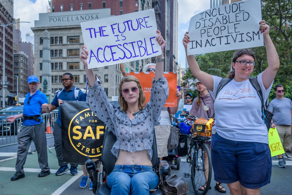 Wheelchair-using person holds up sign that says “The Future Is Accessible,” next to another standing person who is holding an “Include Disabled People In Activism” sign during the fifth annual New York Disability Pride parade on July 14, 2019