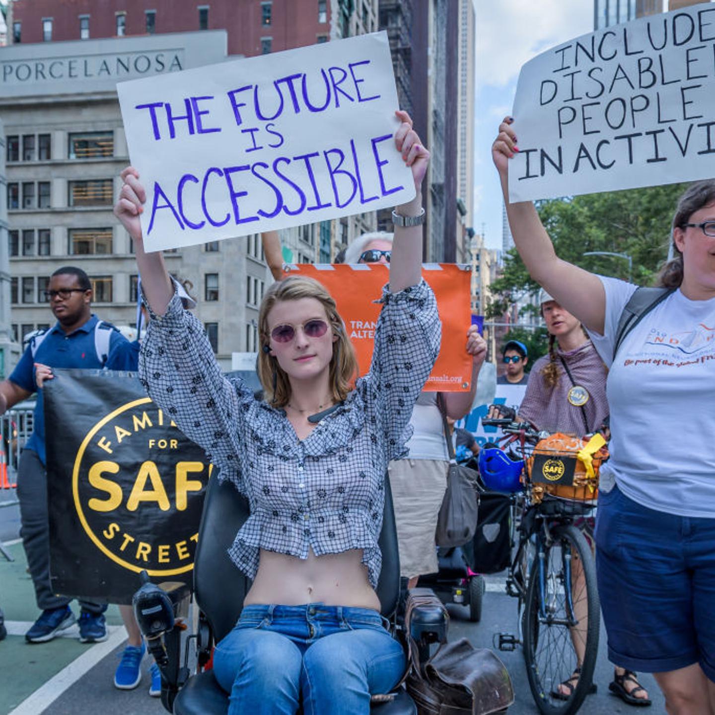 Wheelchair-using person holds up sign that says “The Future Is Accessible,” next to another standing person who is holding an “Include Disabled People In Activism” sign during the fifth annual New York Disability Pride parade on July 14, 2019