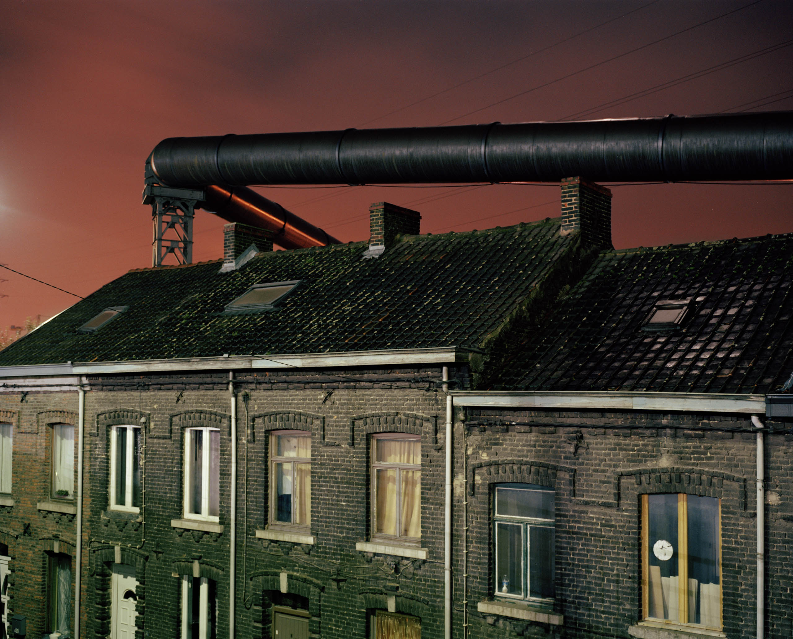 The gas supply tubes run along the houses built near the steel factories of Charleroi. Before the electric upgrade of the blast furnace these tubes used to provide the energy necessary to this operation. The factory and furnace infringe and loom over the