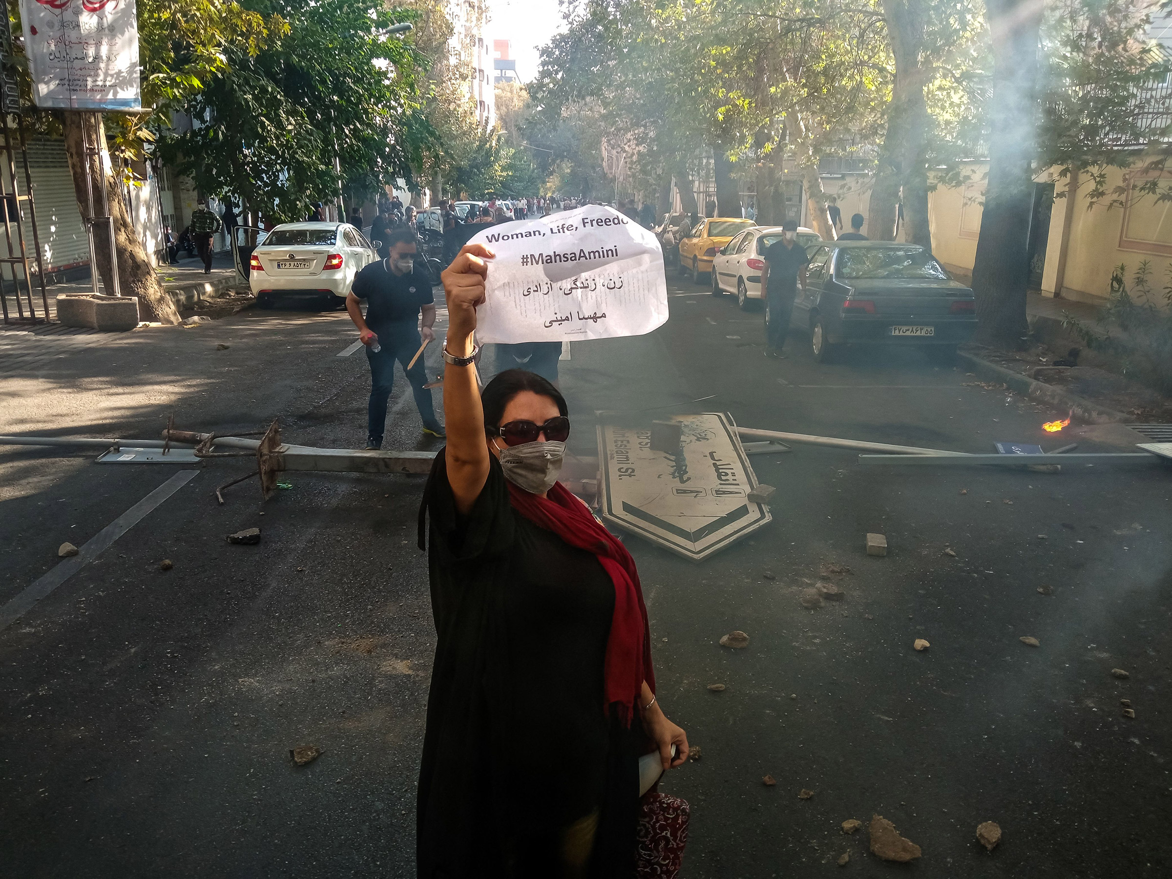 An Iranian woman protester wearing a mask but no hijab holds up a paper reading "Woman, Life, Freedom" and "Mahsa Amini" in front of a street sign on the ground with Revolution and Islamic Republic names on it near Enghelab (Revolution) Street in Tehran,