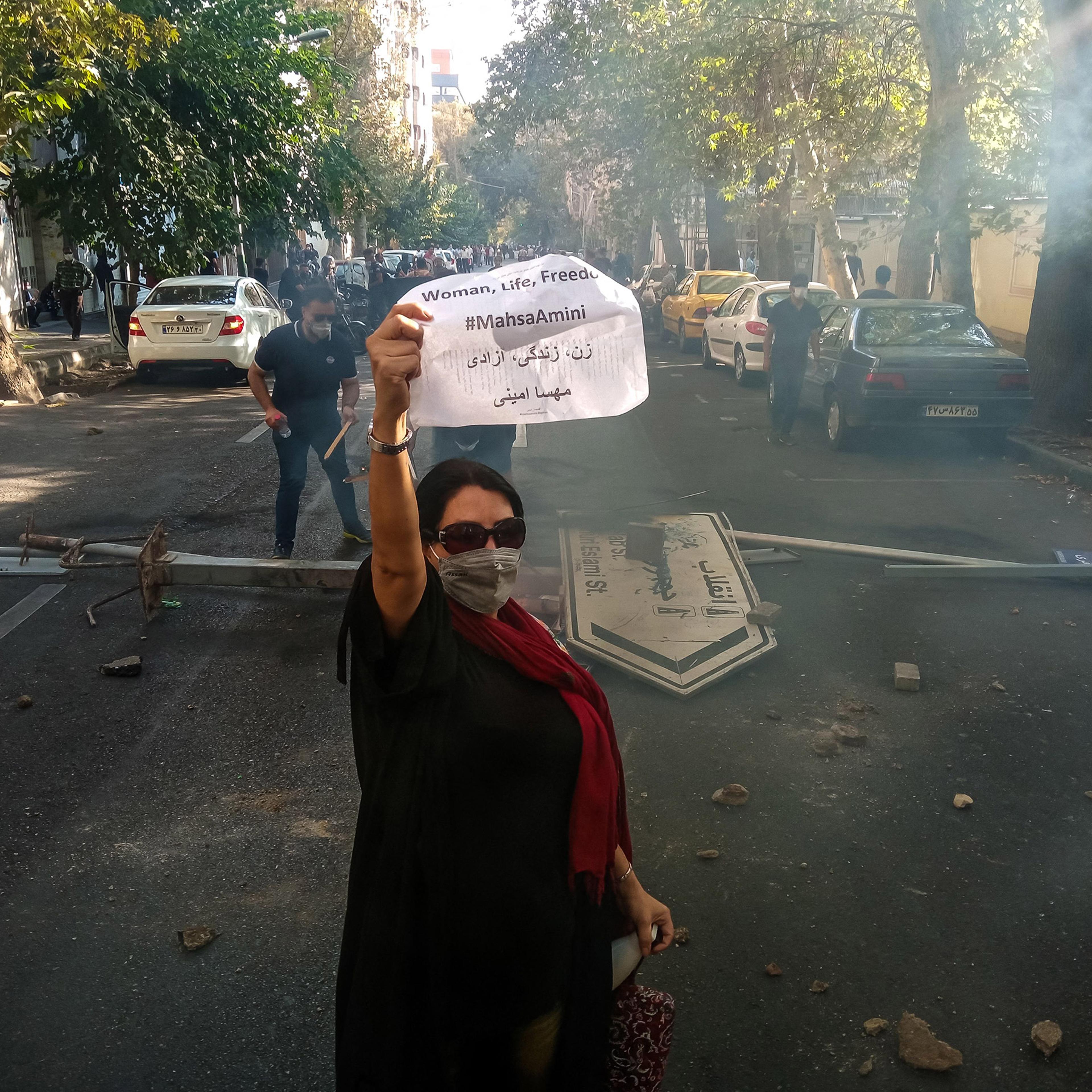 An Iranian woman protester wearing a mask but no hijab holds up a paper reading "Woman, Life, Freedom" and "Mahsa Amini" in front of a street sign on the ground with Revolution and Islamic Republic names on it near Enghelab (Revolution) Street in Tehran,