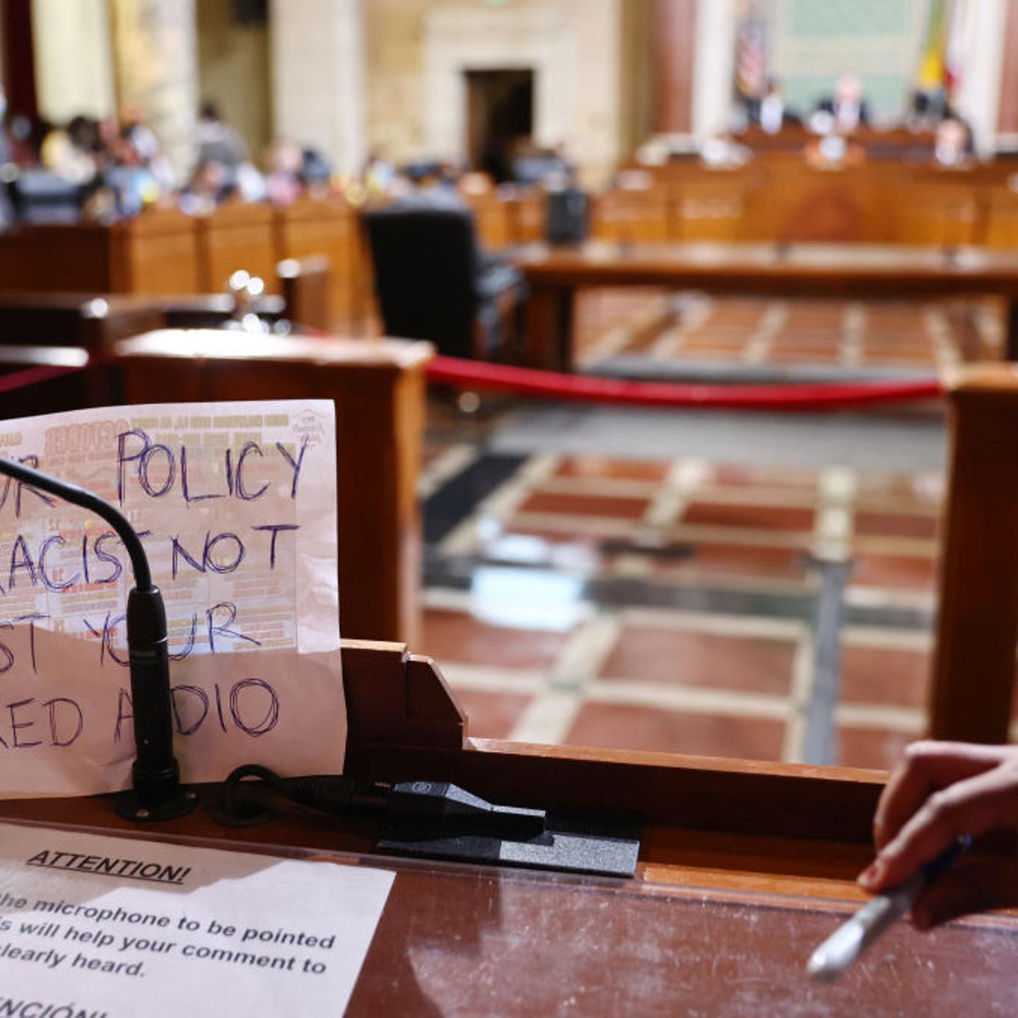 A sign rests against the microphone stand that says “Your Policy Is Racist Not Just Your Leaked Audio,” as protestors demonstrate at the L.A. City Council.