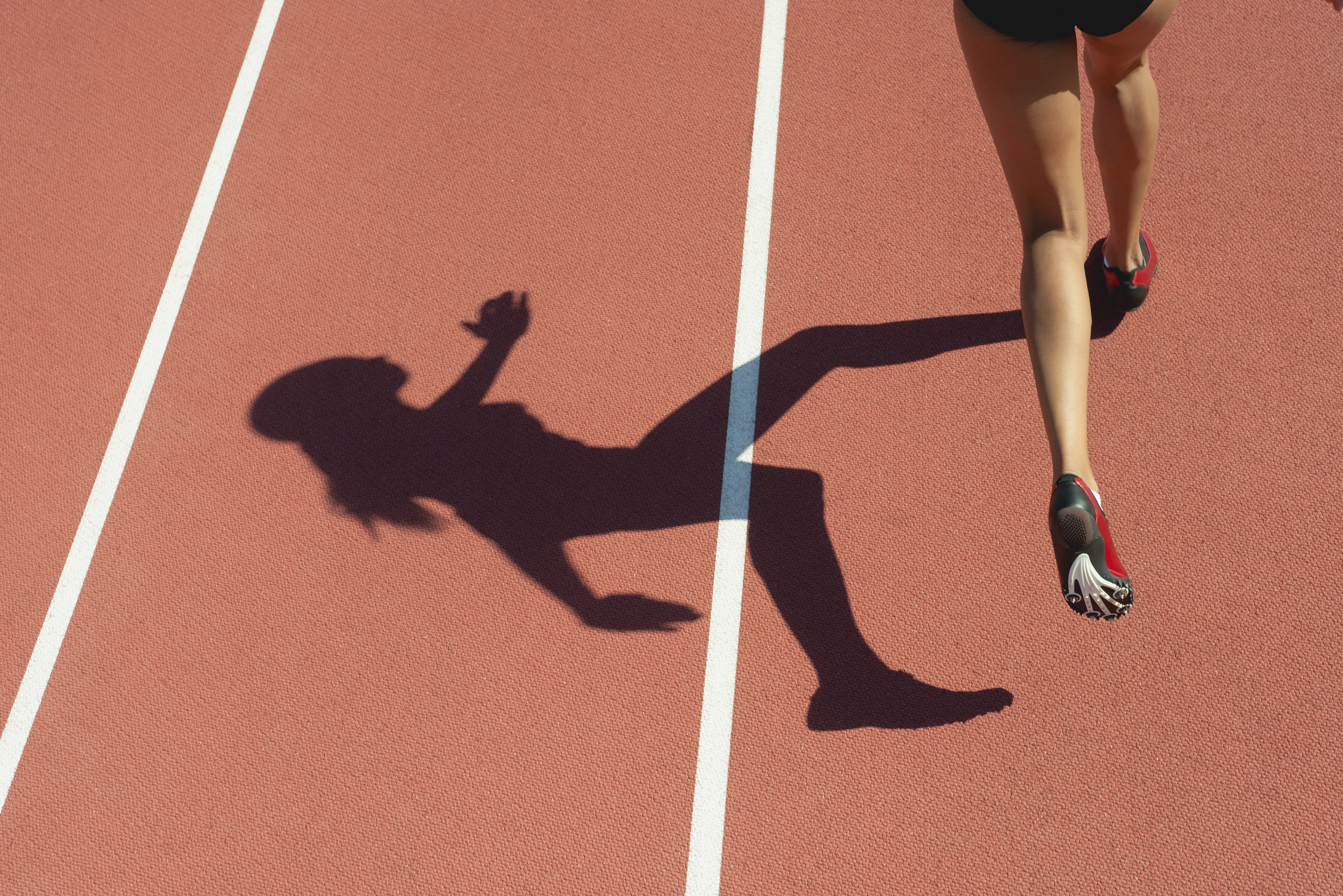Female athlete running on track, low section, focus on shadow