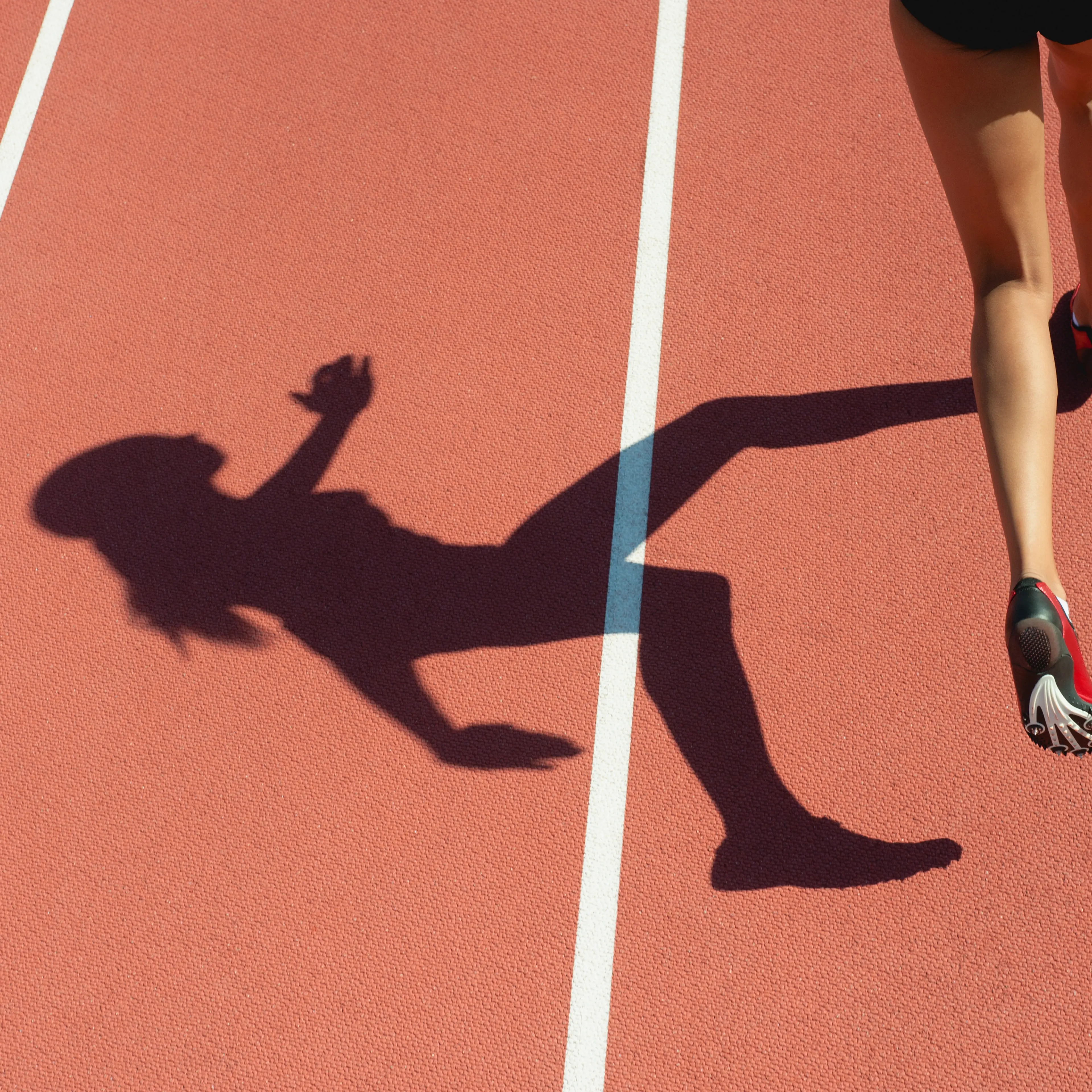 Female athlete running on track, low section, focus on shadow